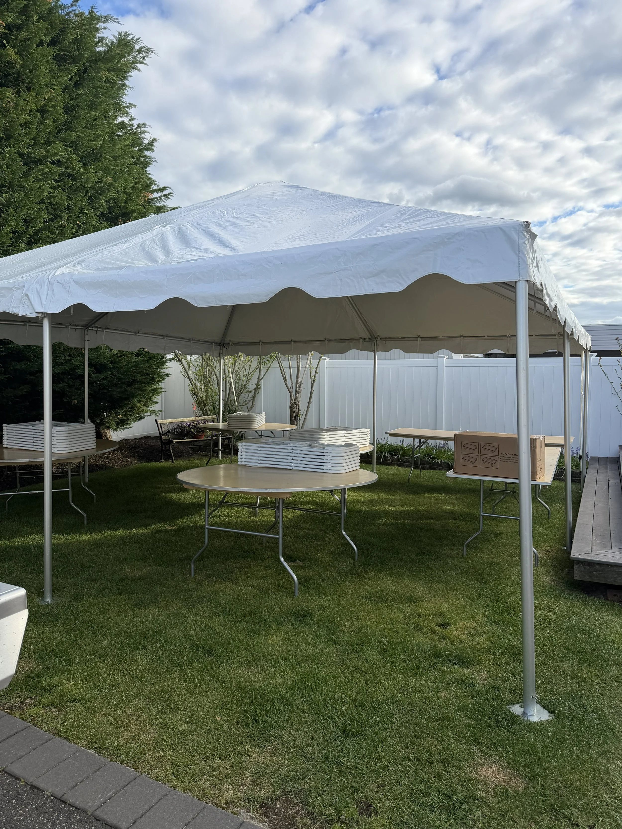 Empty white canopy tent set up on a grassy lawn, with folded tables and chairs underneath, surrounded by a white fence and greenery, under a partly cloudy sky.