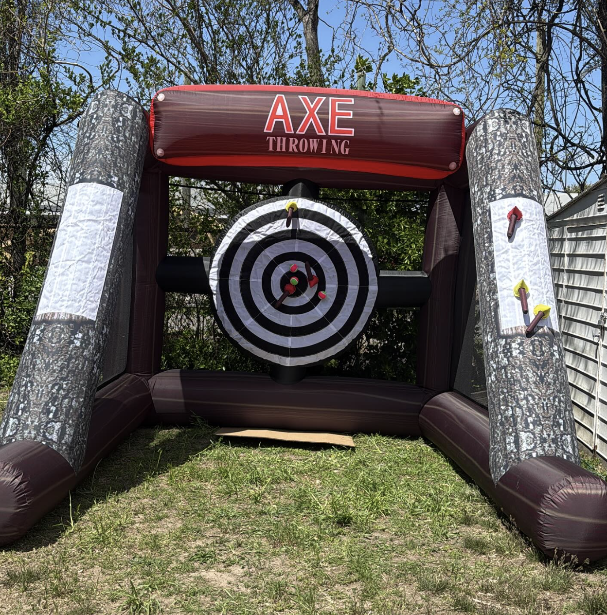 Inflatable outdoor axe throwing game with a target in the center and two axe props on the sides, set up on grass with trees and a fence in the background.