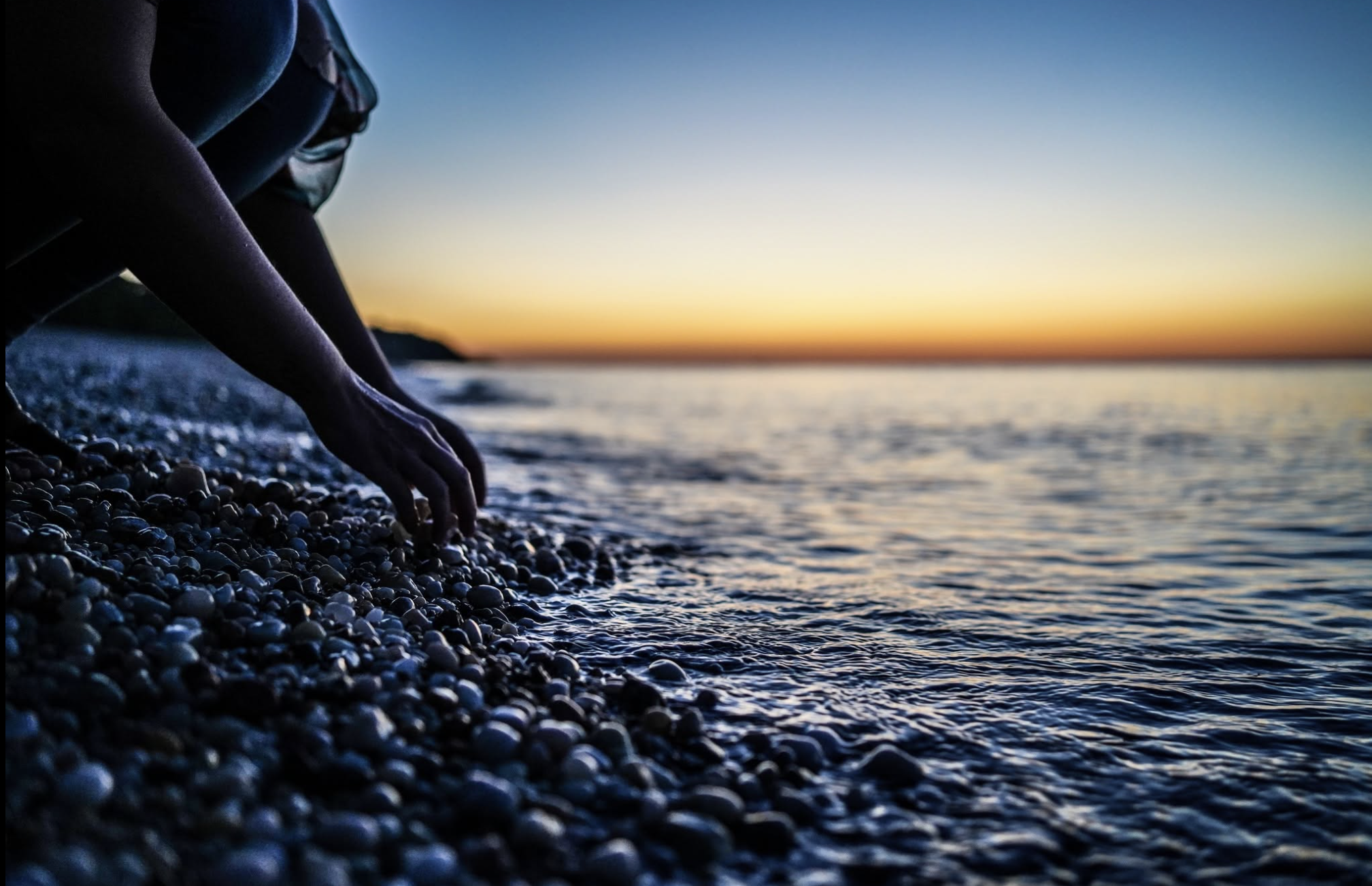 Person crouching on a pebble beach during sunset, reaching into the water.