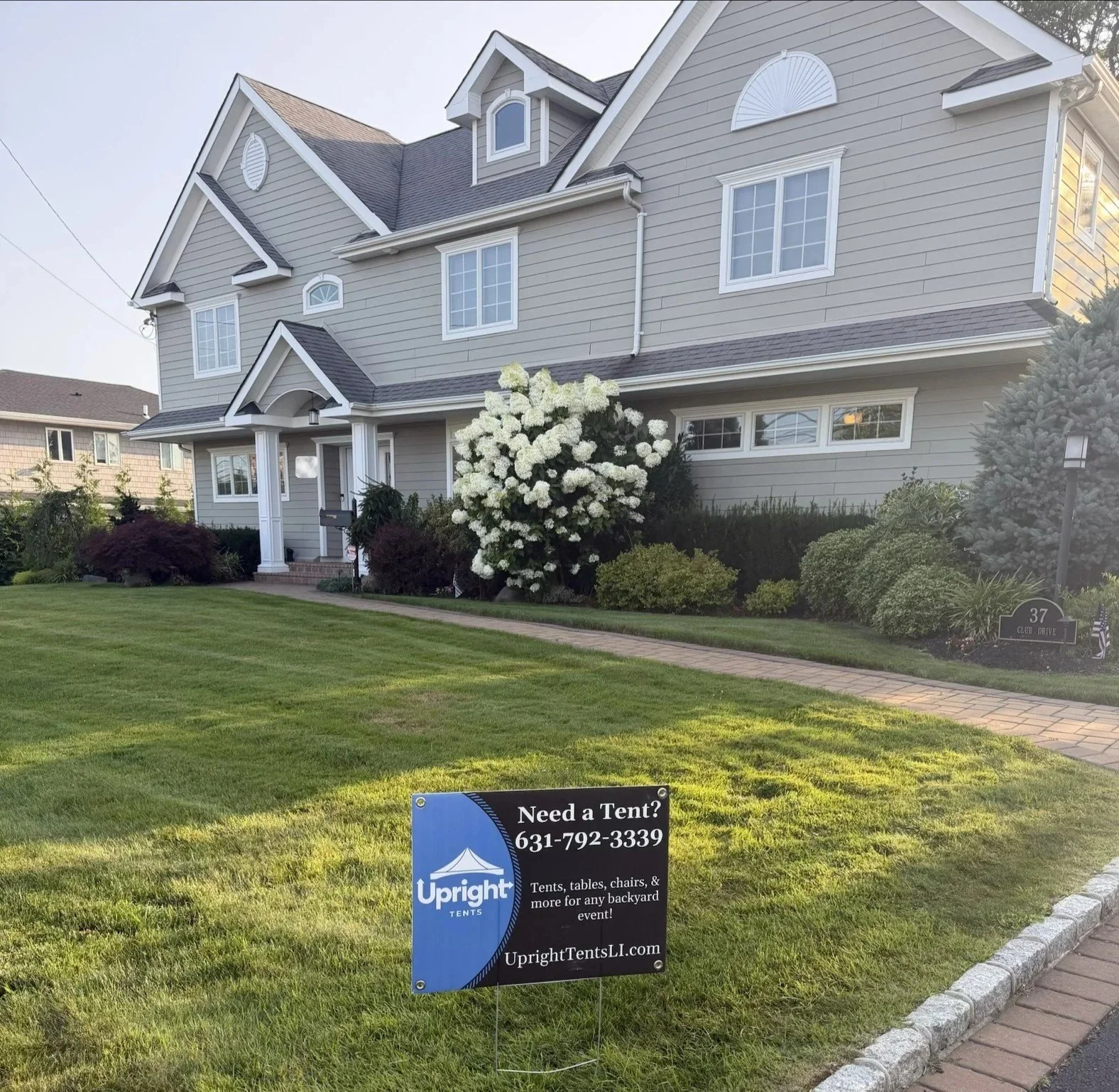 A two-story house with grey siding, white trim, and multiple gable roofs. There is a flower bush with white flowers and a well-maintained lawn in front. A sign on the lawn advertises tent rentals, with the text 'Need a Tent? 631-792-3339 UbrightTentsLL.com'.