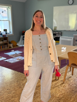 Person in a classroom wearing a light-colored jumpsuit and cardigan, holding a pink item in hand, with tables, chairs, and educational materials in the background.