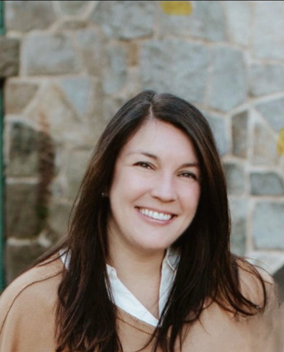 A woman with long dark hair smiling in front of a stone wall, wearing a white shirt and tan sweater.