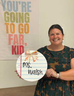 A smiling woman holds a decorated circular sign with the words "Ms. Walsh" written in a script font. Behind her, a colorful poster reads "YOU'RE GOING TO GO FAR, KID." The woman wears a dark patterned dress and stands against a plain wall.