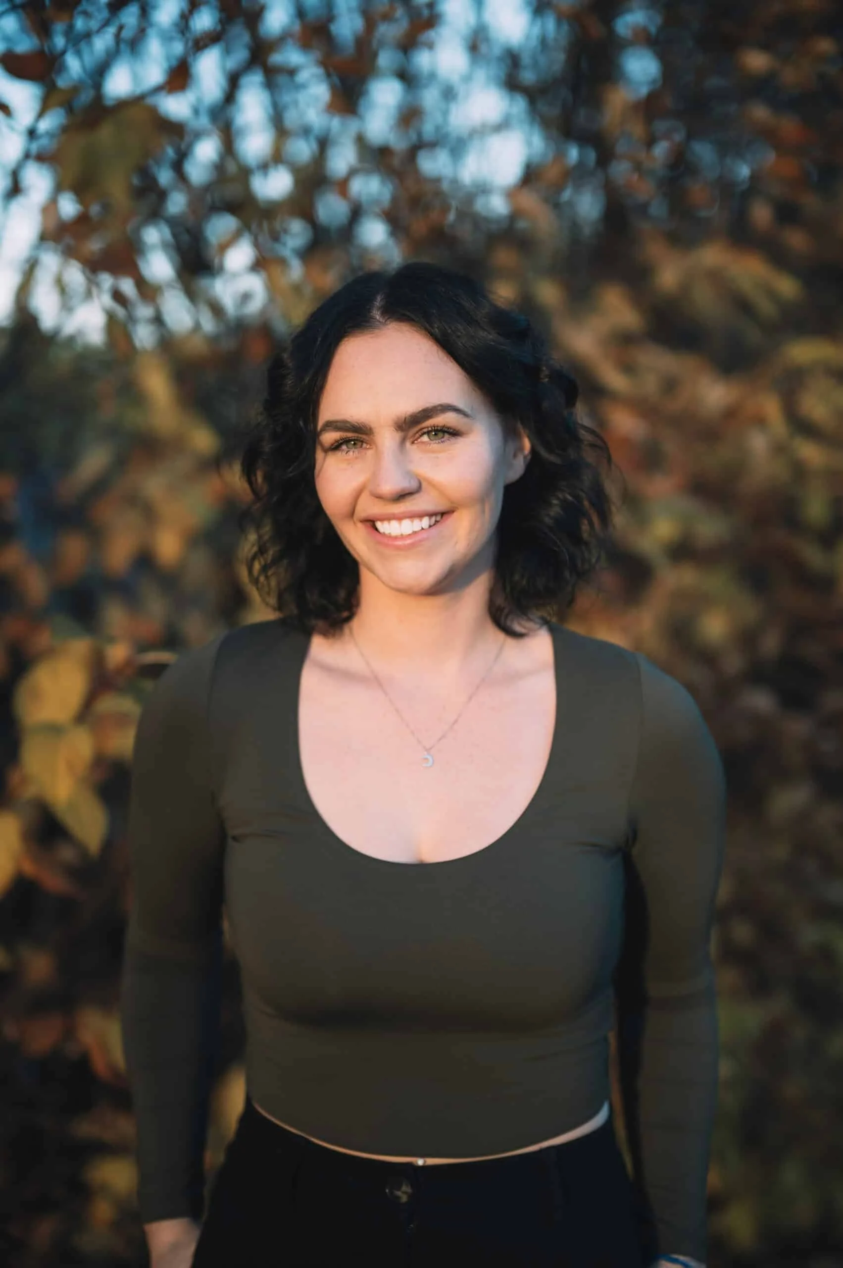 A young woman with black curly hair, smiling and wearing a dark green long-sleeve top, standing outdoors in front of autumn-colored foliage.