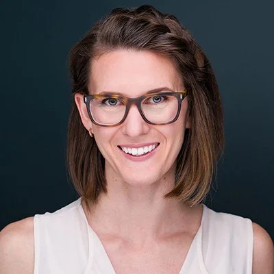 A woman with shoulder-length brown hair, wearing glasses and a white sleeveless top, smiling against a dark background.