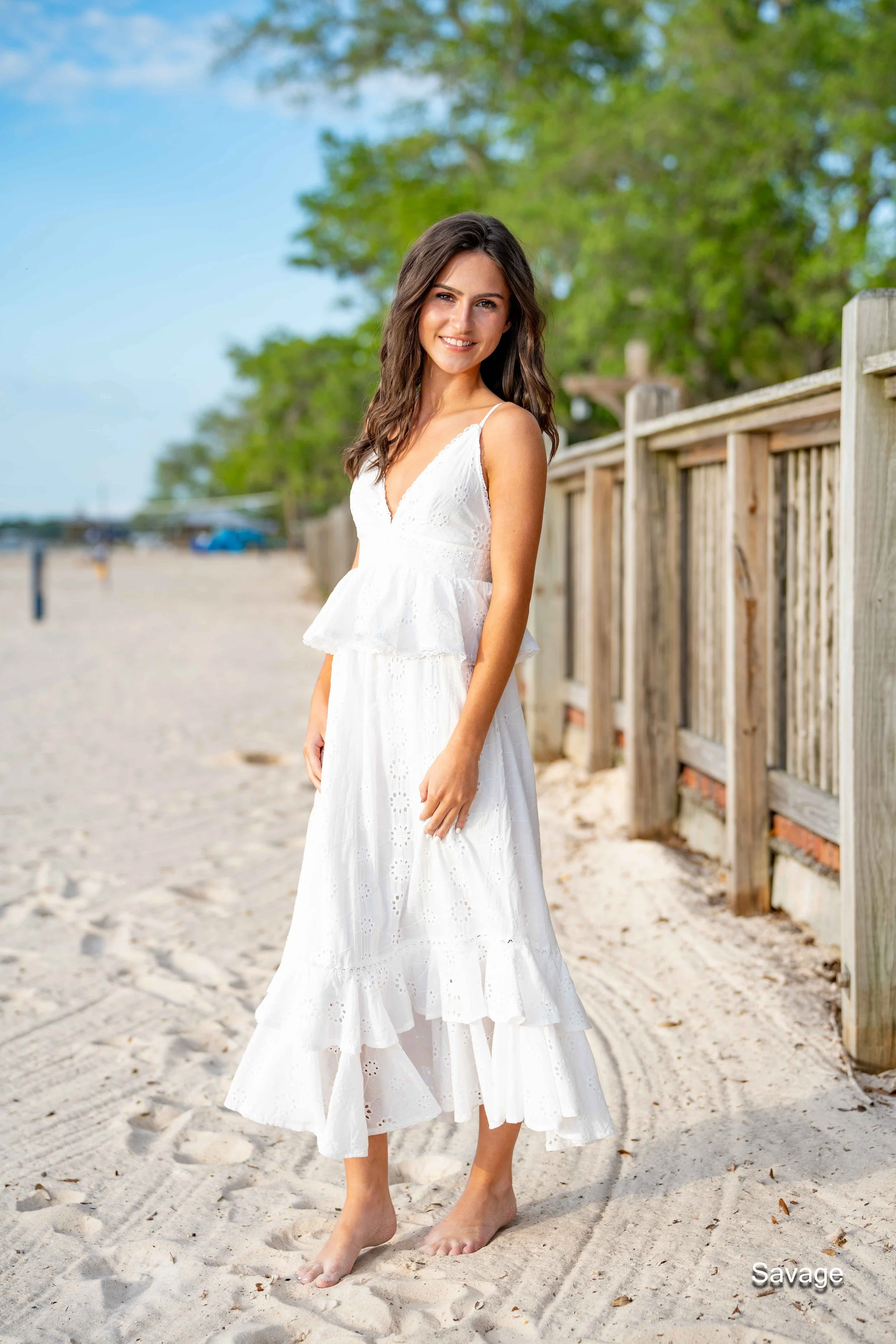 Woman in a white summer dress standing barefoot on a sandy beach with a wooden fence and trees in the background.