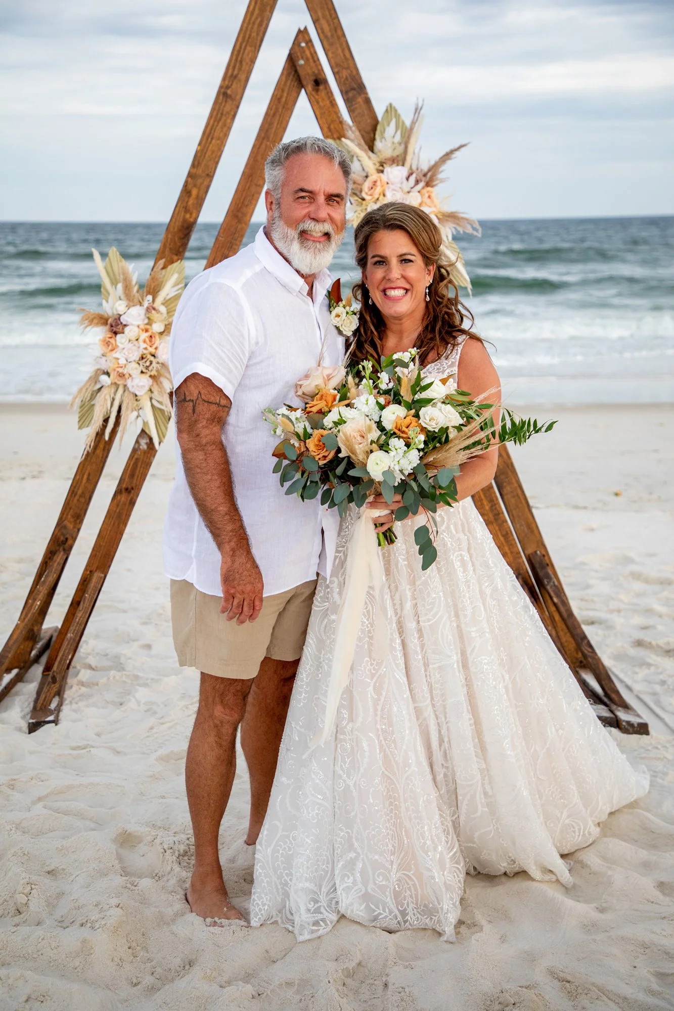 A couple standing on a beach in wedding attire. The woman is wearing a white lace dress and holding a bouquet of flowers, while the man is in a white shirt and khaki shorts. They stand in front of a wooden triangular arch decorated with flowers.