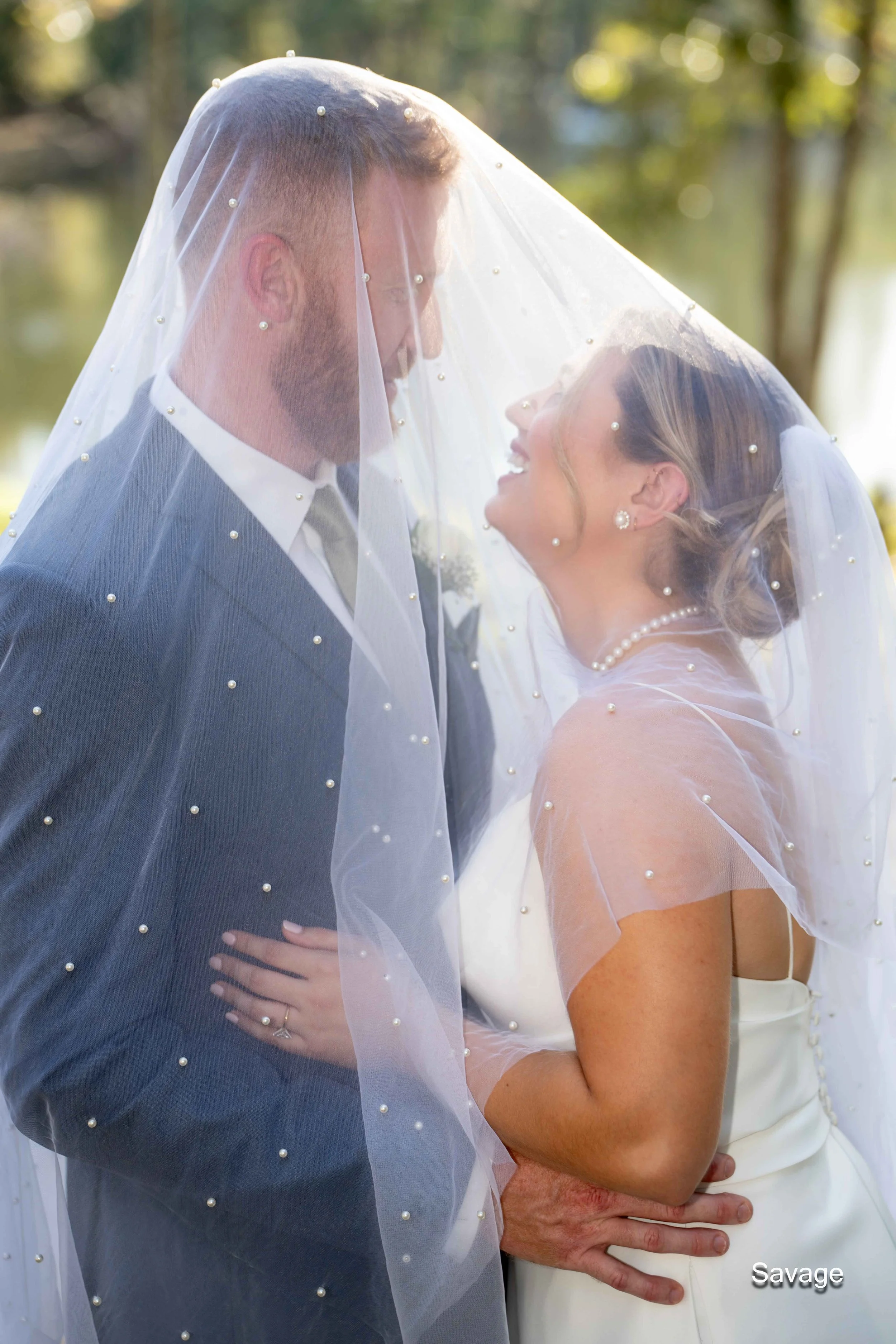 Bride and groom under a pearl-studded veil, smiling at each other in an outdoor setting.