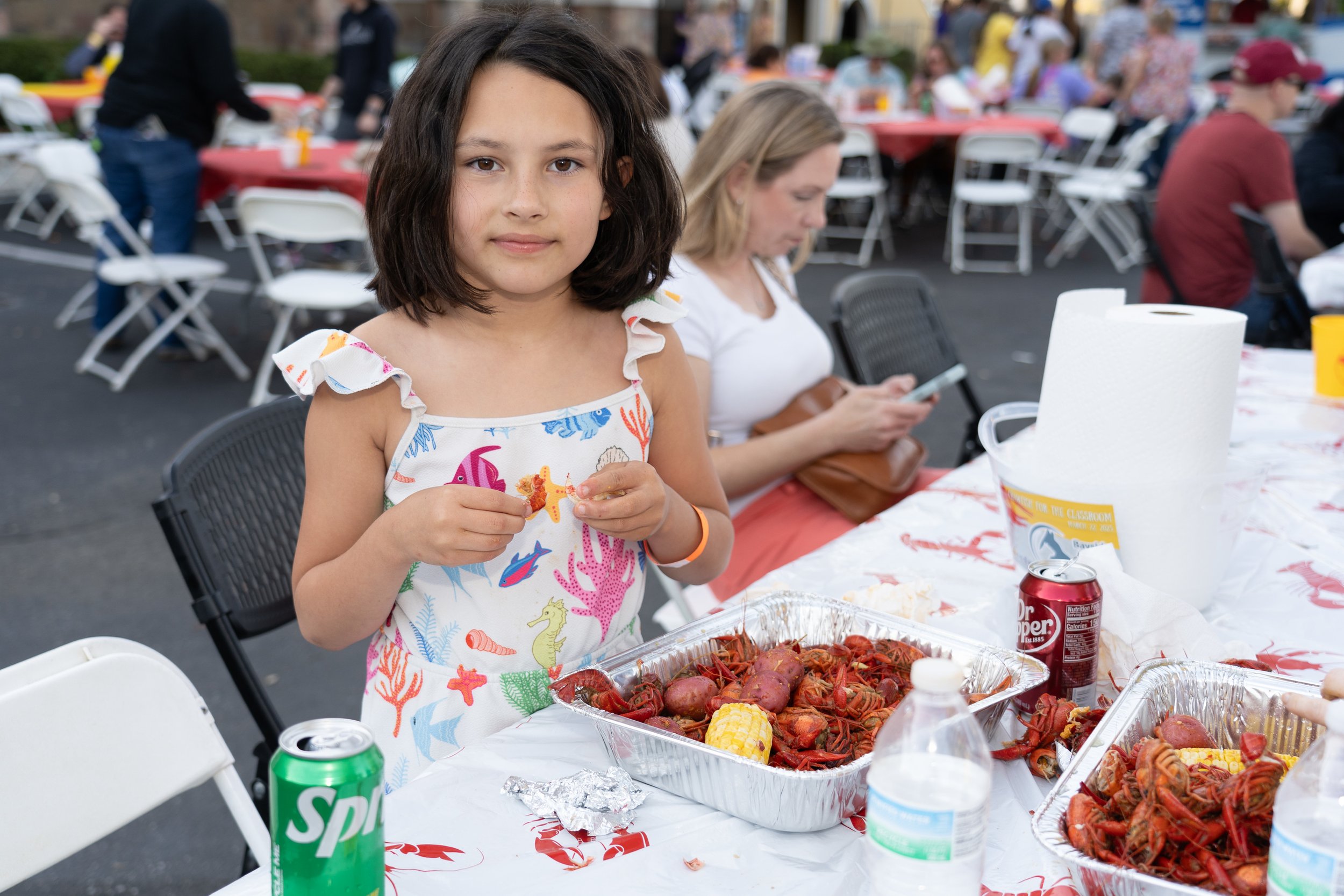 Girl in a colorful dress at a crawfish boil holding food, with trays of crawfish, potatoes, and corn on the table. People sitting at tables in the background.