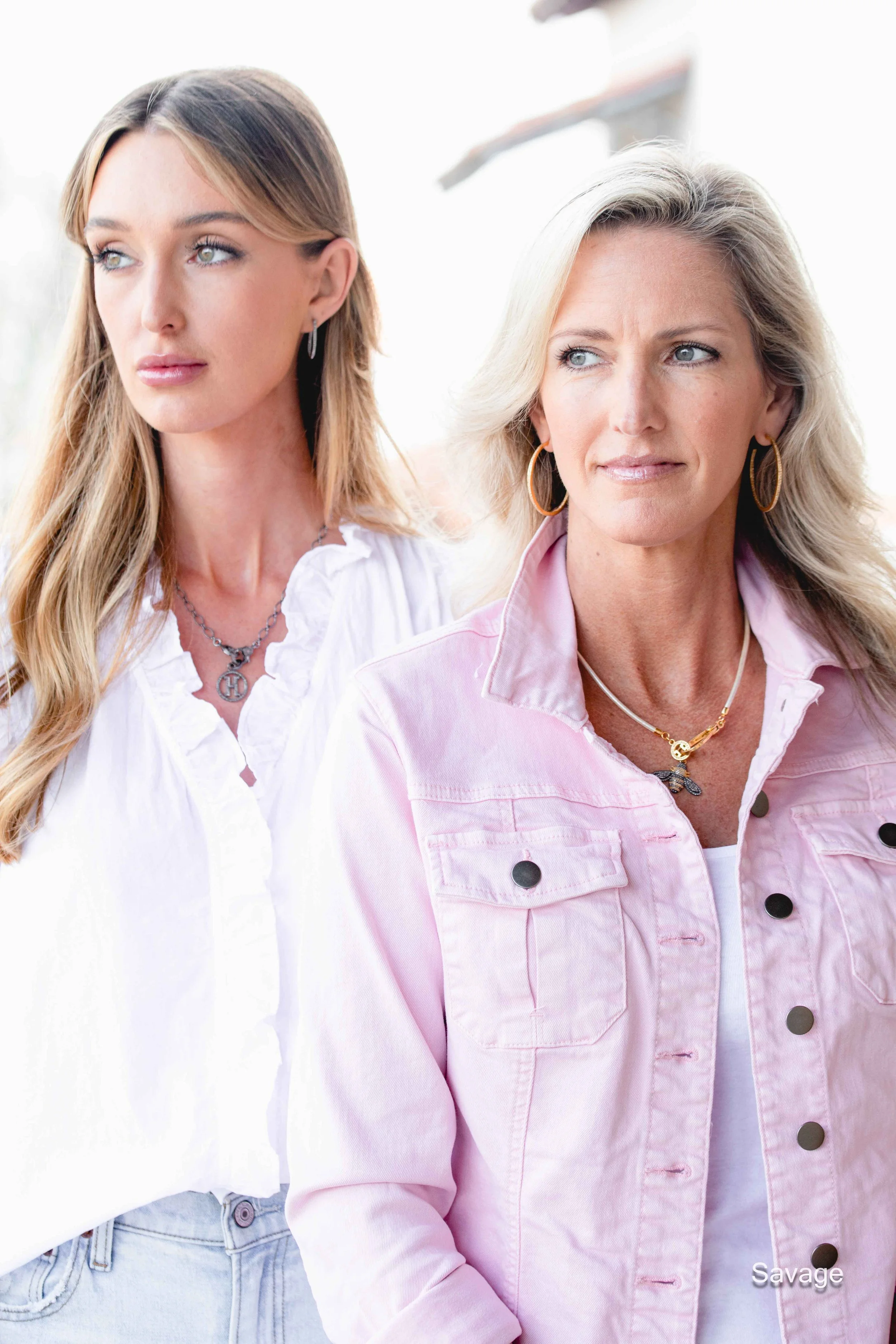 Two women with long hair, one wearing a white blouse and the other a pink jacket, standing outdoors.