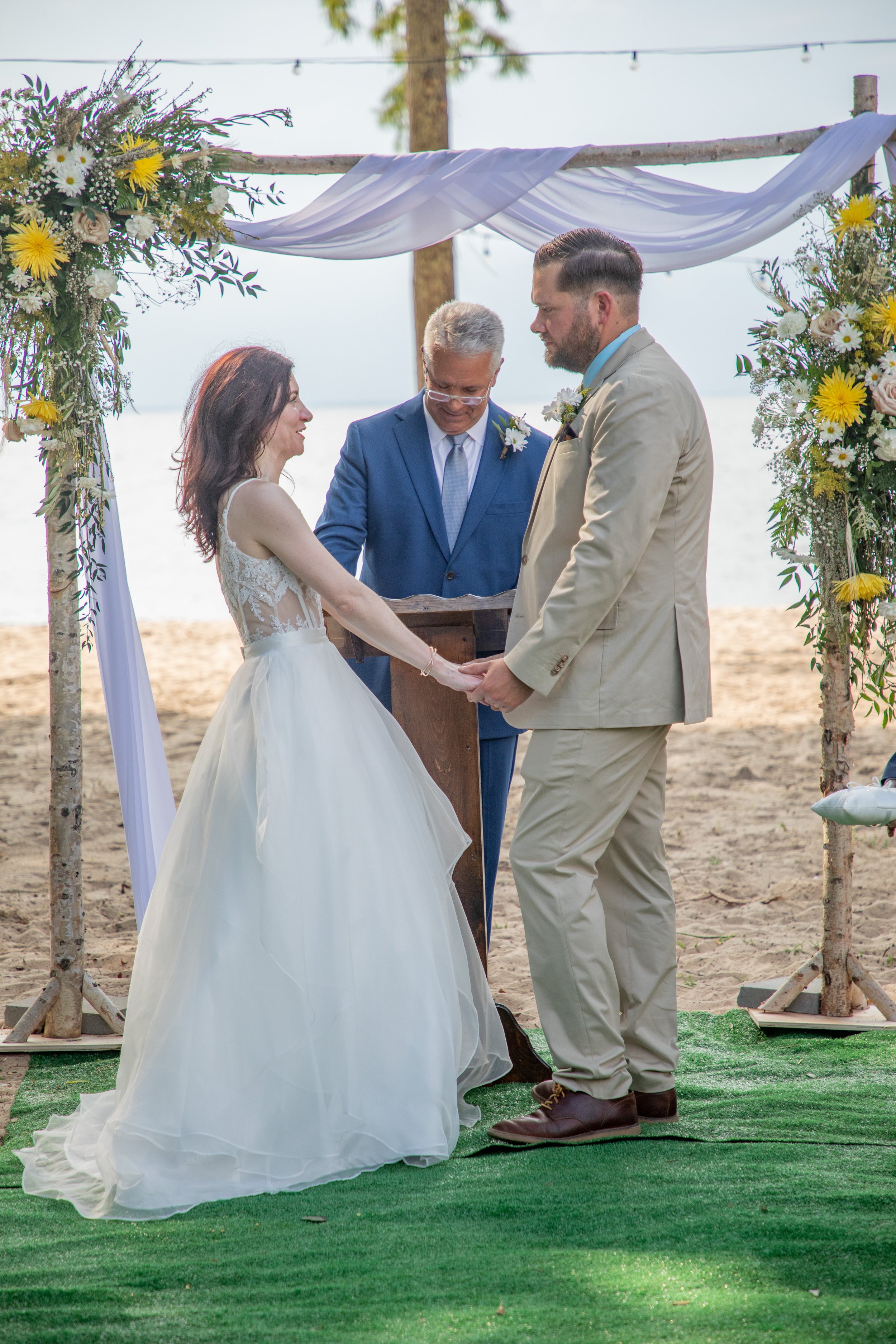 A couple holding hands during a beach wedding ceremony with an officiant in a blue suit, standing under a floral arch.