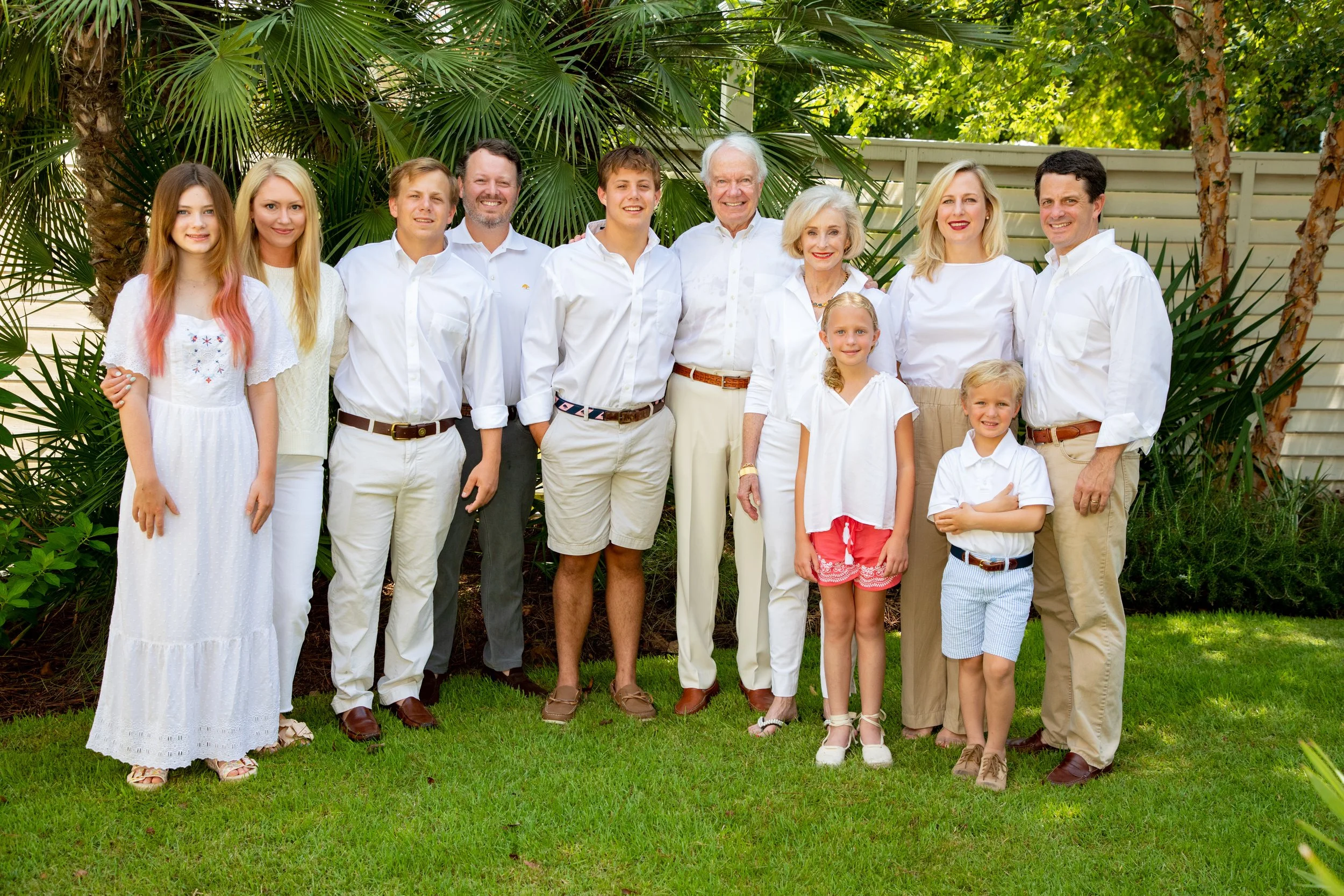 A group of adults and children wearing white clothing posing together outside on a lawn with green foliage in the background. The family appears happy and relaxed.