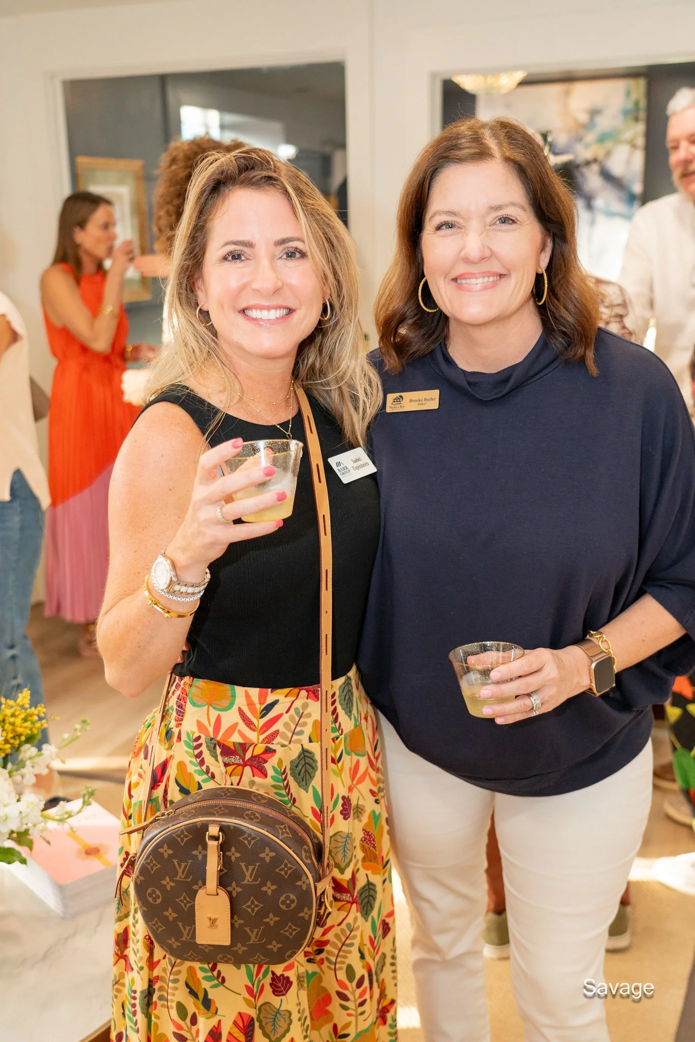 Two women at an event holding drinks, one wearing a black top and colorful skirt with a Louis Vuitton bag, the other in a navy shirt with a name tag. People and decorations are visible in the background.