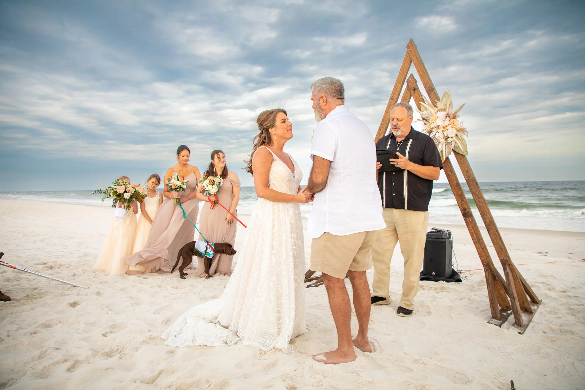 Beach wedding ceremony with bride and groom exchanging vows, officiant reading from a book, bridesmaids holding bouquets, and a dog on a leash, under a wooden triangular arch.
