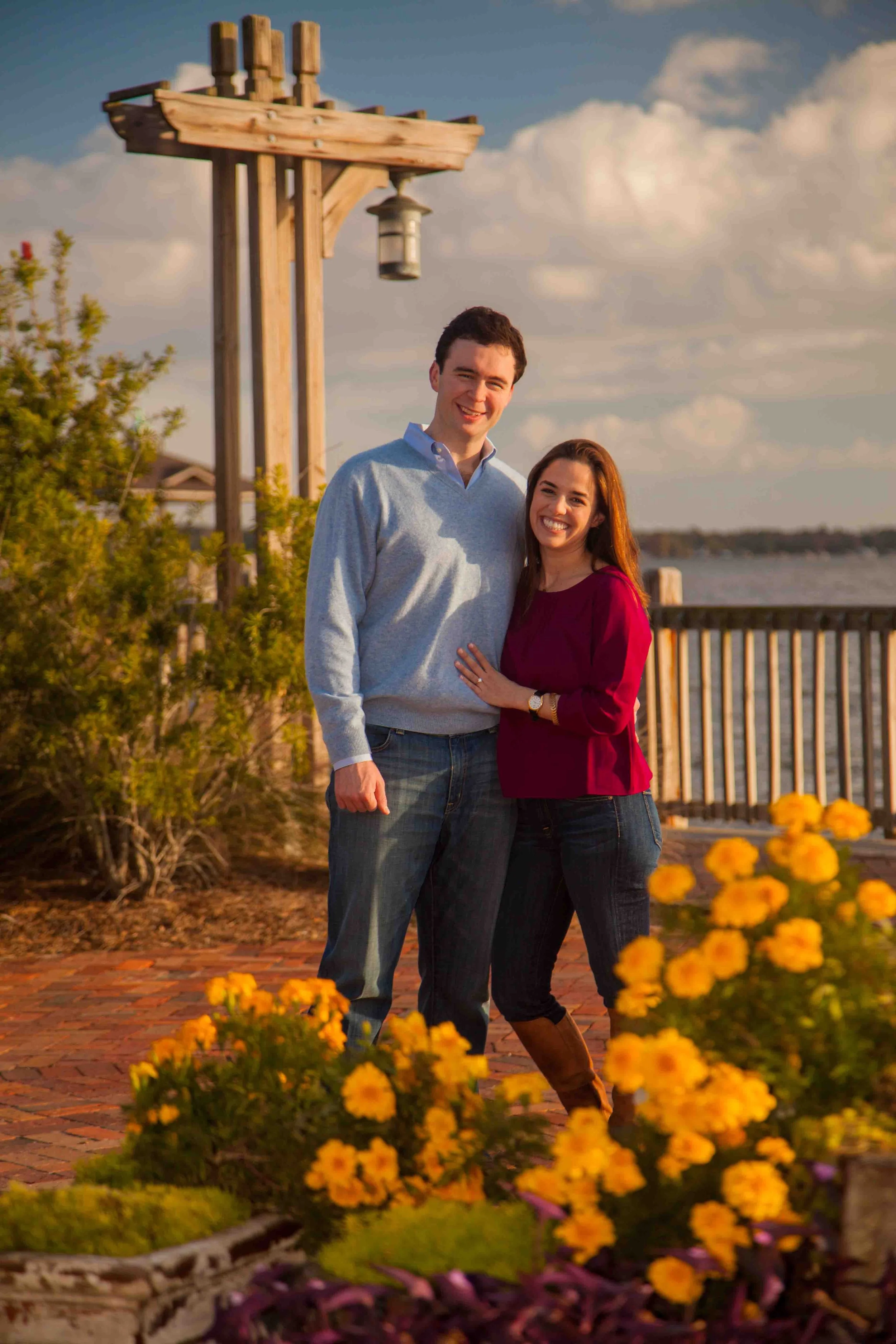 Couple posing for a photo in a garden with yellow flowers, a wooden pergola, and a river or lake in the background.