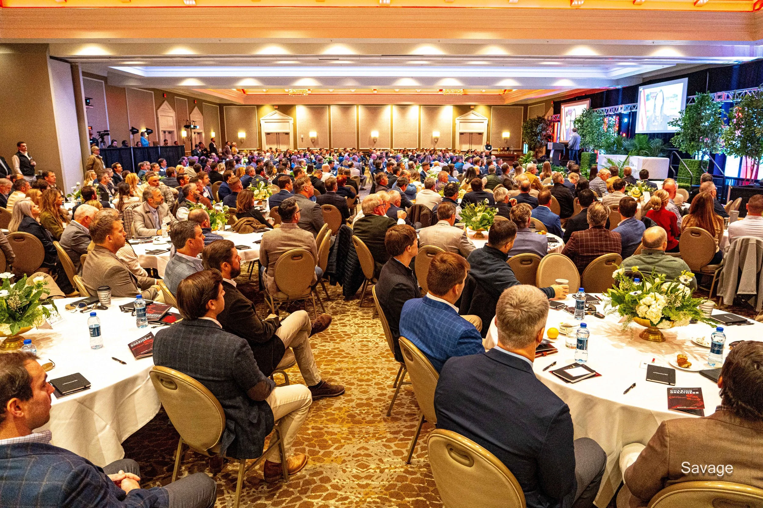 Large conference room filled with attendees sitting at tables facing a stage with a presenter, decorated with plants and flowers.
