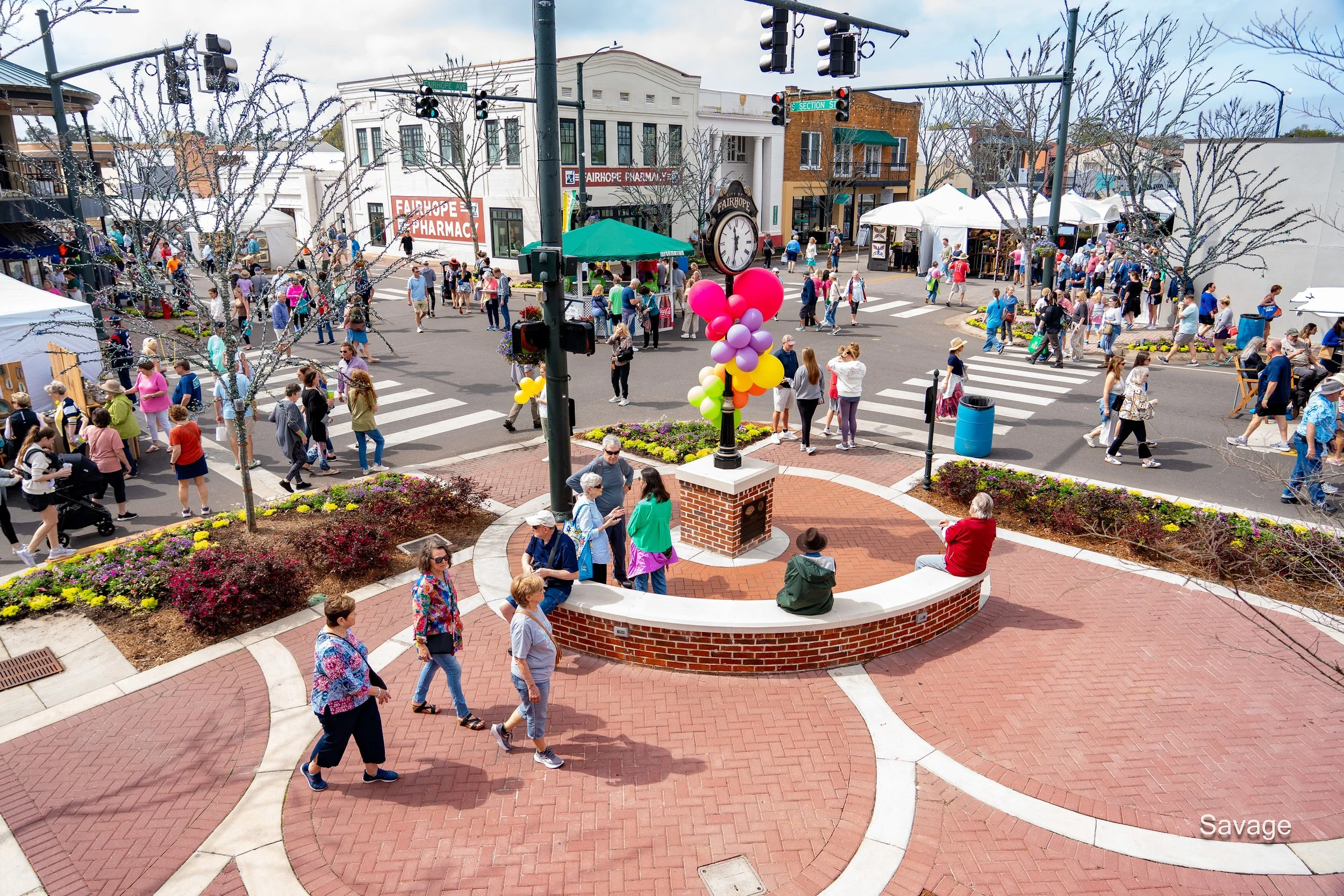 A lively street fair with people walking along a brick-paved area. Multicolored balloons are tied to a post with a clock. Tents and booths are visible in the background, with a mix of attendees enjoying the event.