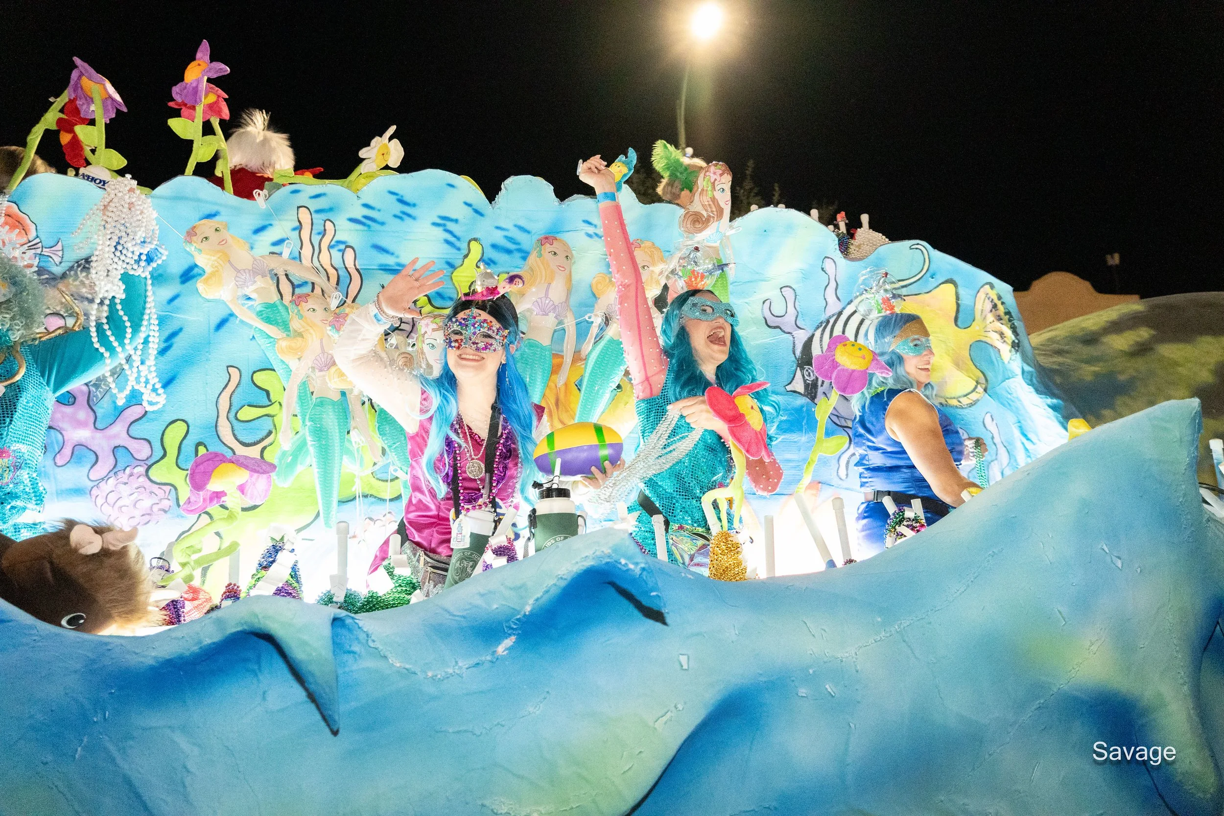 People in colorful costumes and masks on a festive parade float with an under-the-sea theme, featuring vibrant blue waves and mermaid decorations.