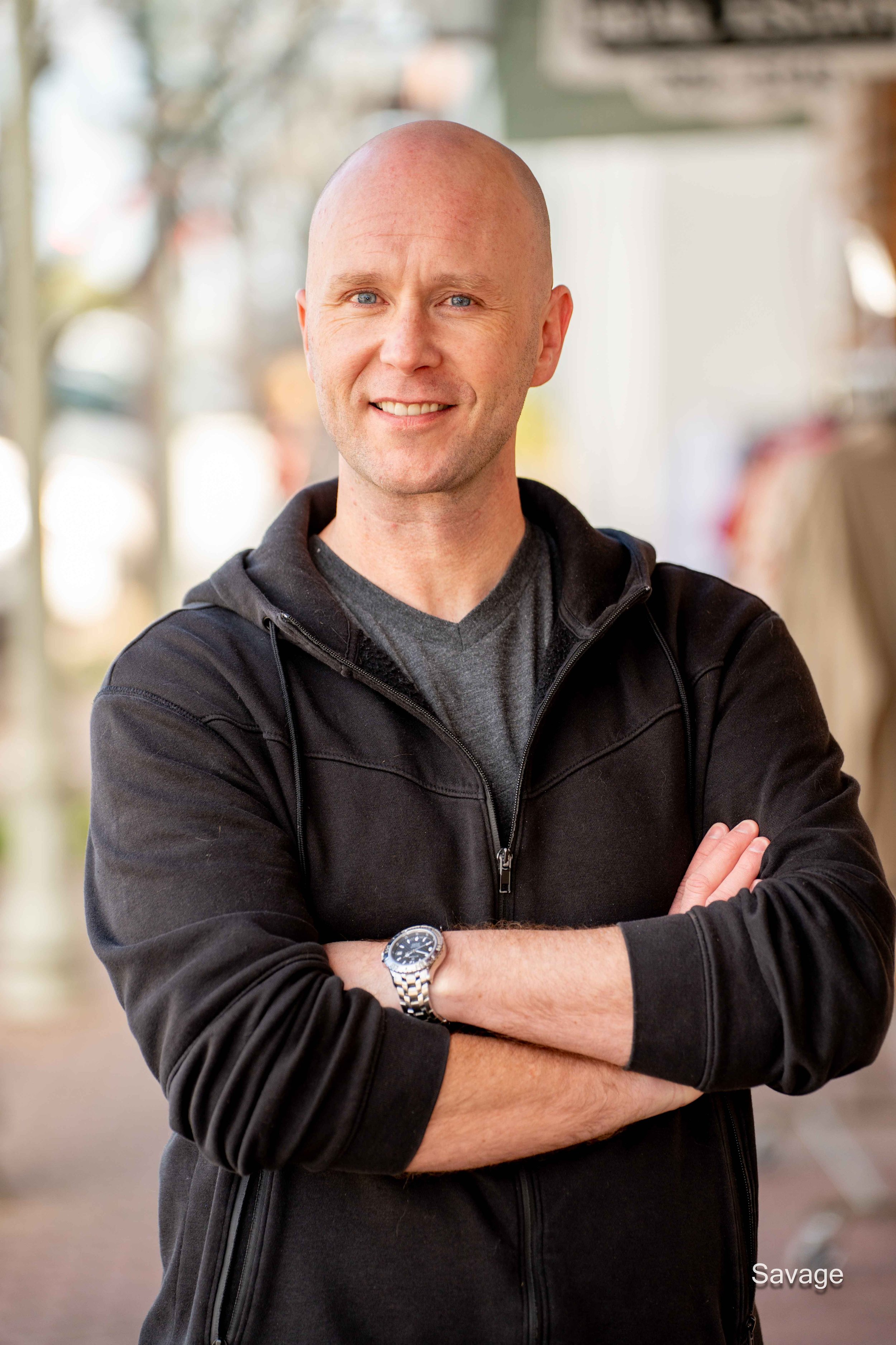 Smiling bald man in a black hoodie with arms crossed, wearing a watch, standing outdoors.