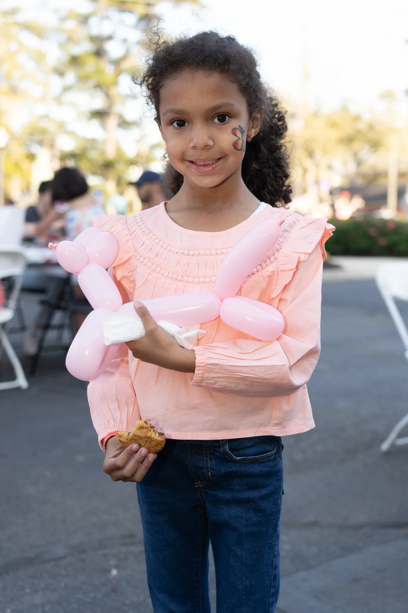 Young girl holding a pink balloon animal and a cookie, with a butterfly face painting, outdoors.