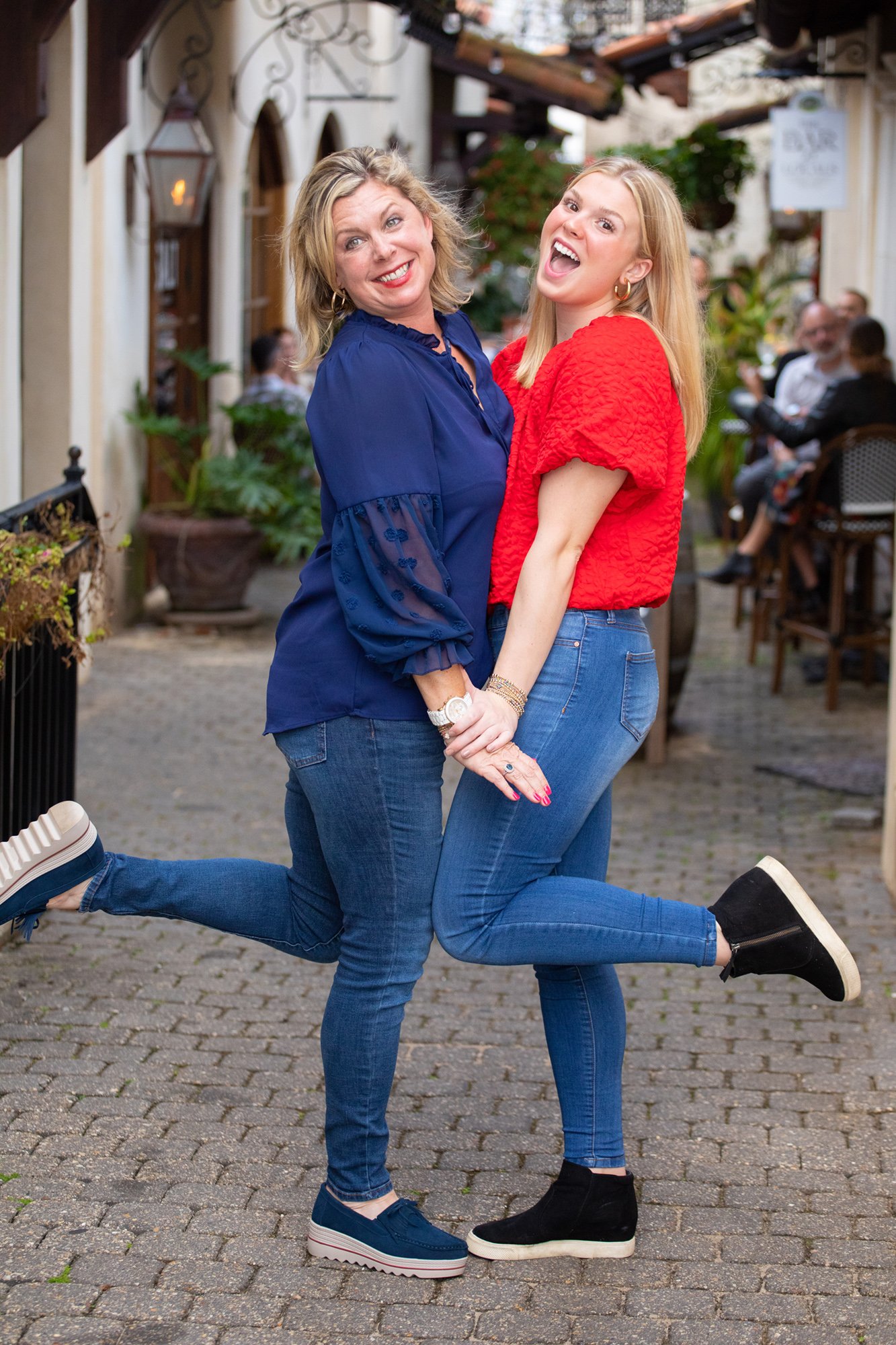 Two women posing playfully on a cobblestone street, wearing blue jeans and colorful tops, smiling and with one leg lifted. Outdoor café in the background.