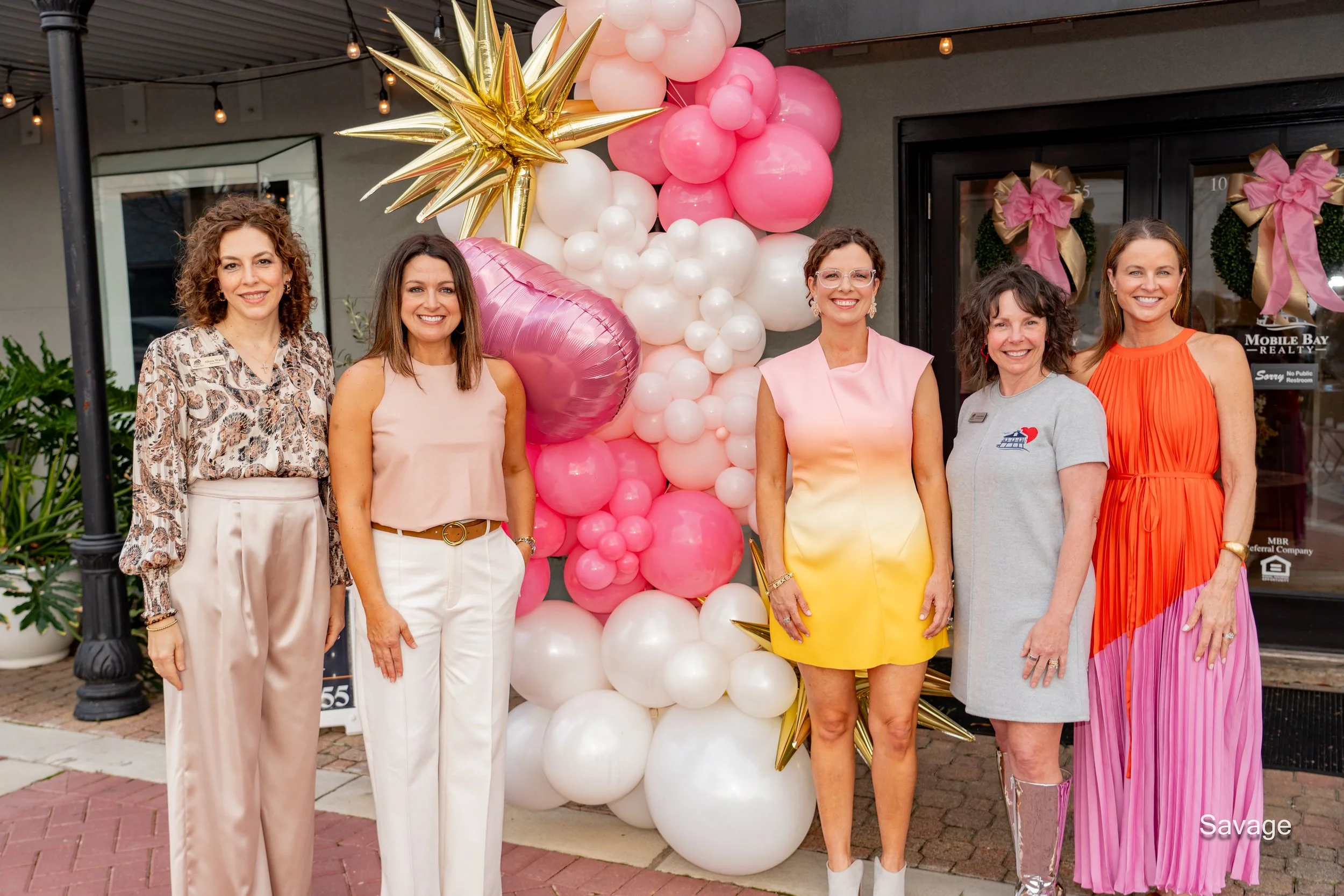 Group of five women standing together, smiling, in front of a balloon arrangement with gold, pink, and white balloons outside a storefront.