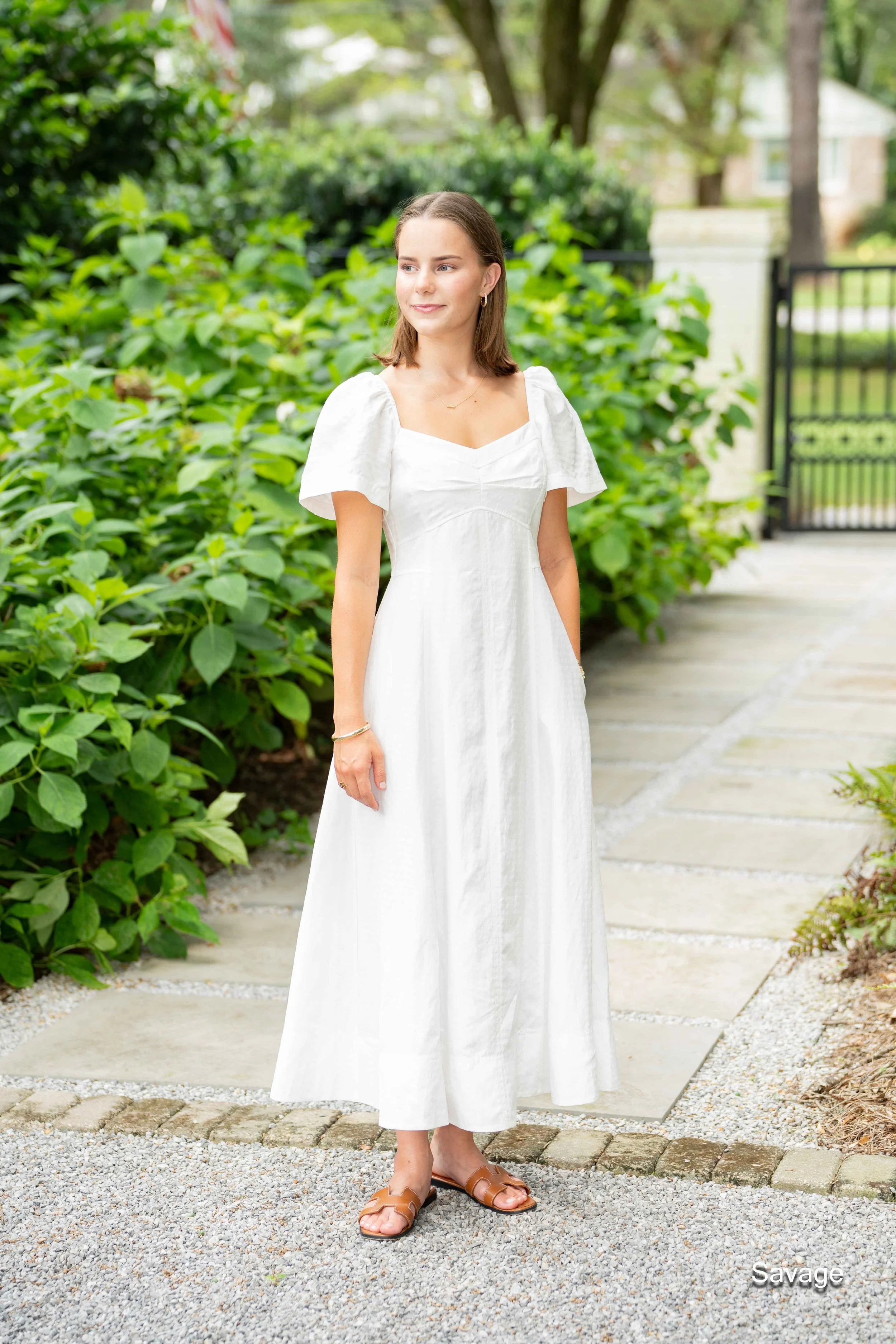 Woman in a white dress standing on a stone path with green foliage background.