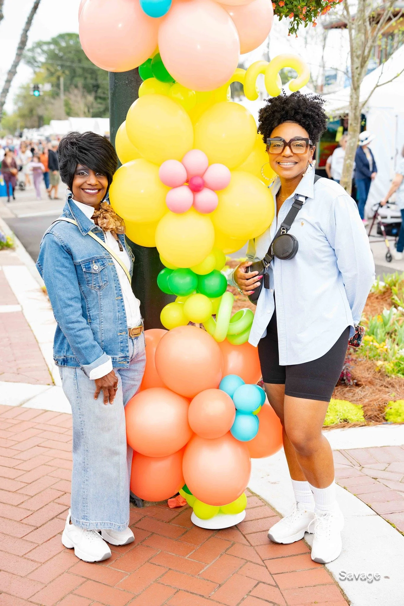 Two women smiling beside a colorful balloon arrangement shaped like flowers at an outdoor event.