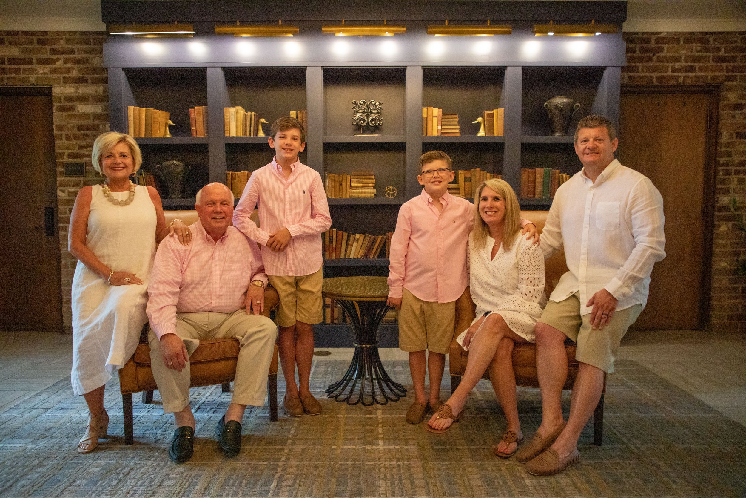 A family of six posing in front of a bookshelf. Two adults are seated, with a woman in a white dress and a man in a pink shirt and light pants. Four others stand, including two boys in matching pink shirts and khaki shorts, and two adults in white at