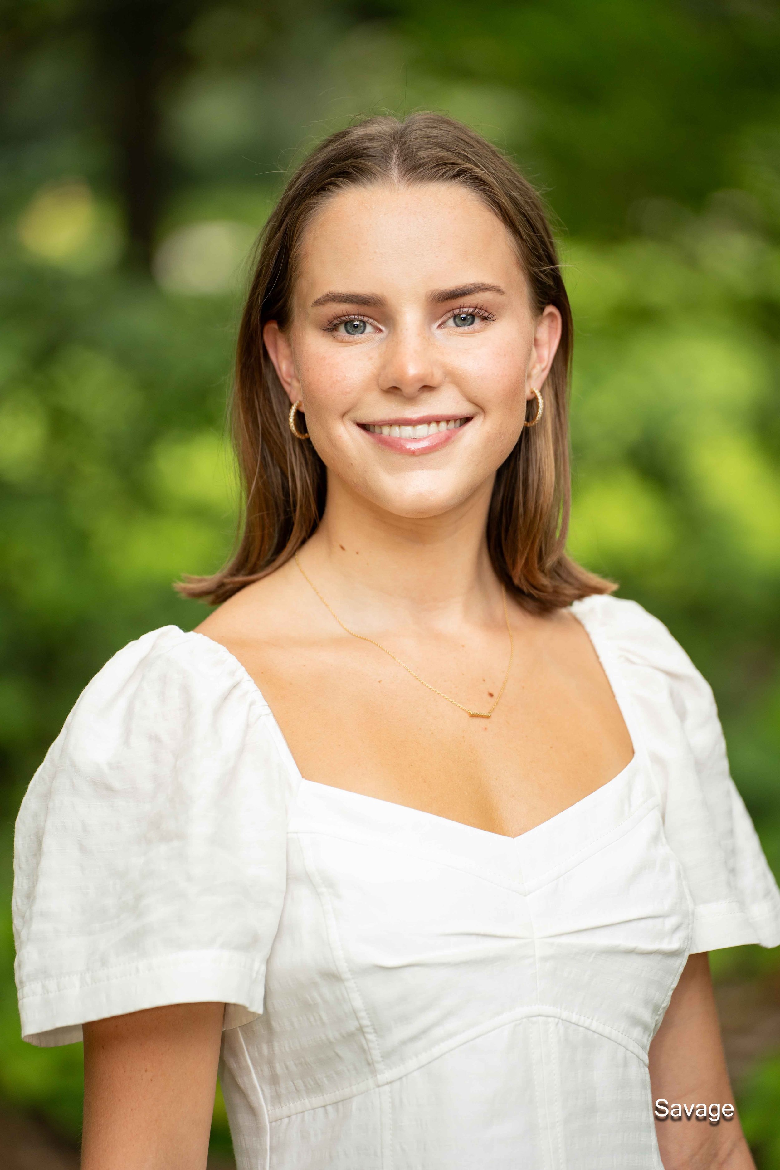 Person smiling in a white dress against a green outdoor background.