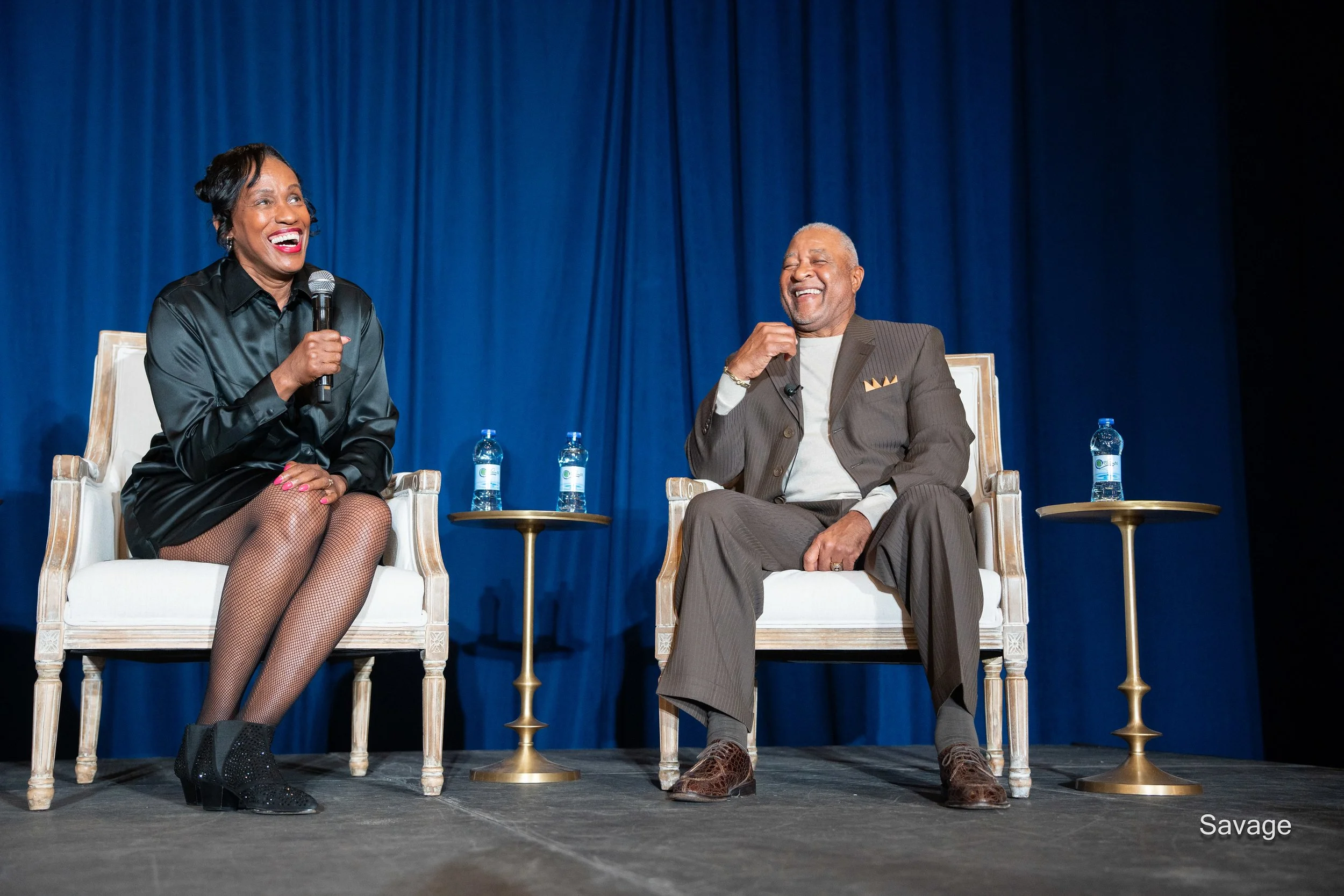 Two people sitting on stage with a blue curtain backdrop, smiling and holding microphones. They are seated on white chairs with small tables holding water bottles beside them.