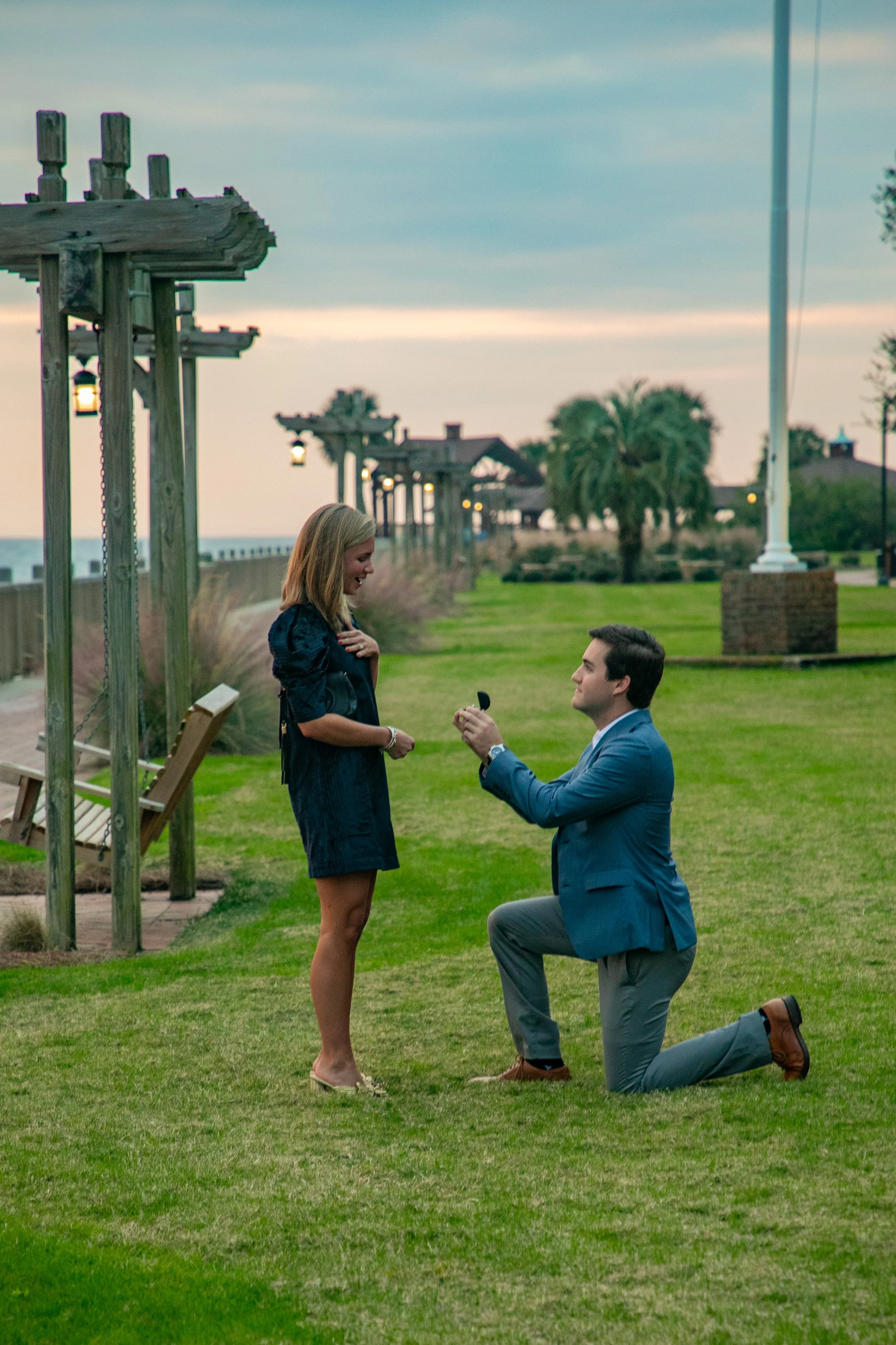 Man proposing to woman on grassy area by the beach, with wooden structures and palm trees in the background.