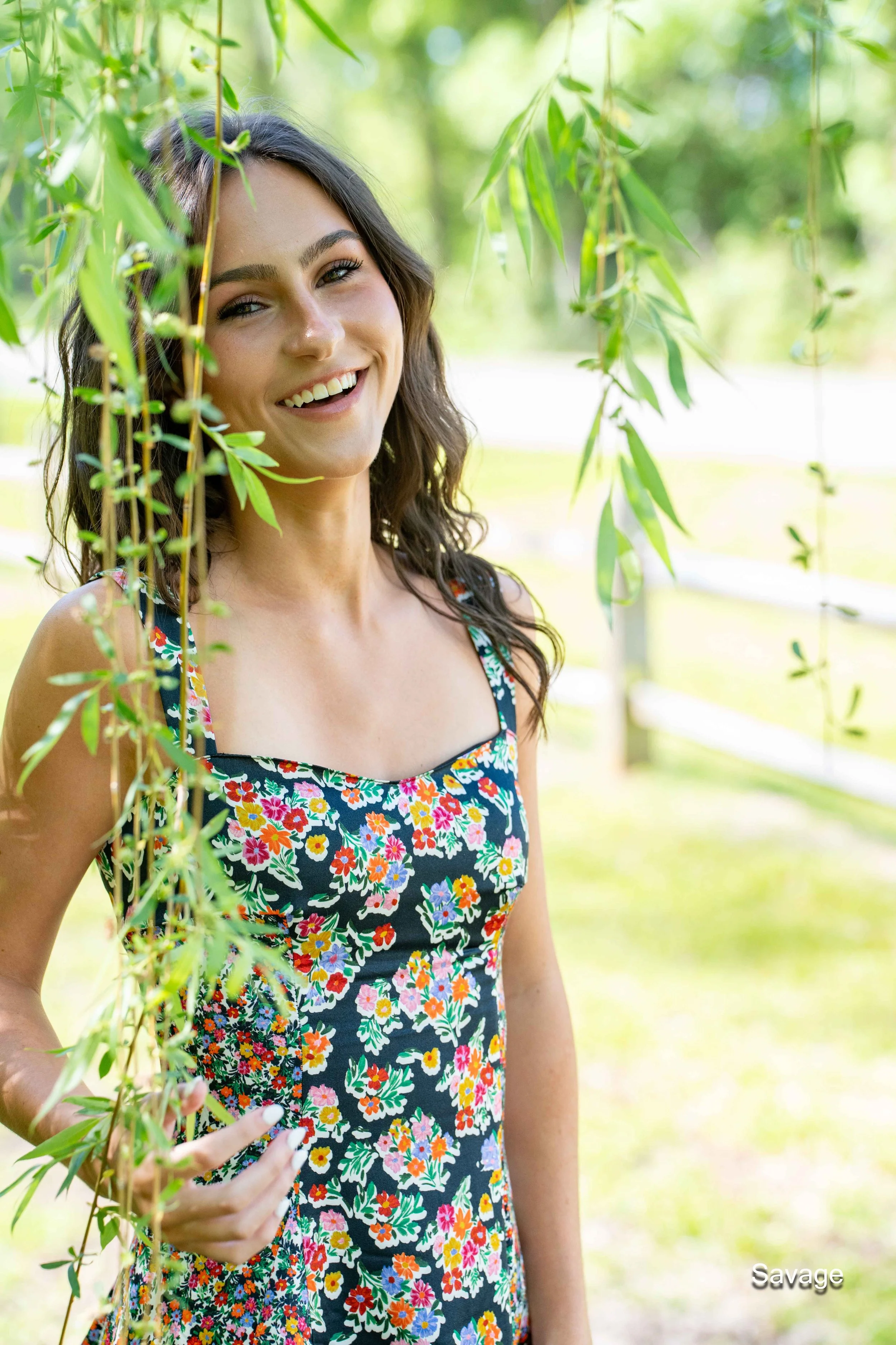 Smiling person in a floral dress standing outdoors near leafy branches.