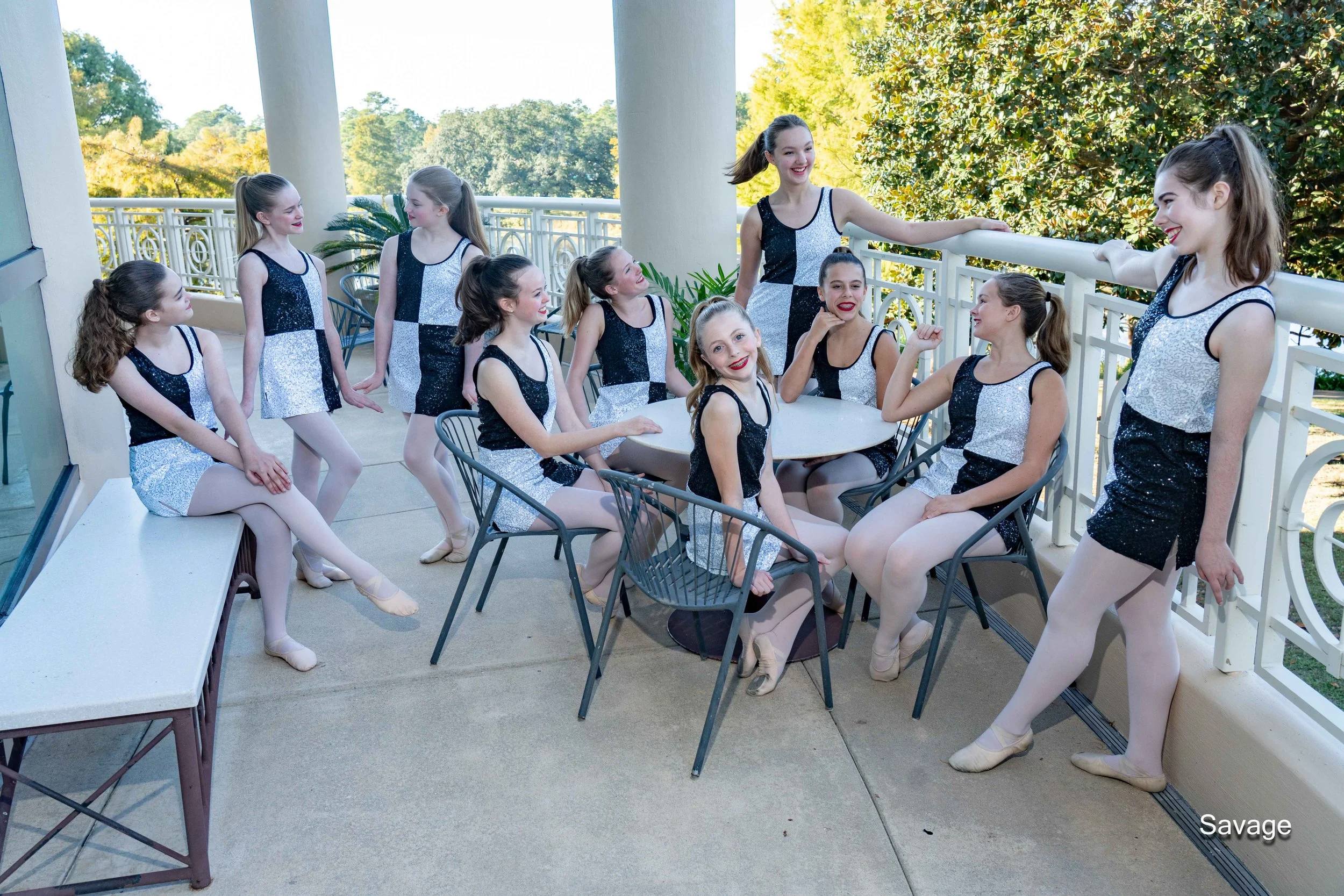 Group of young dancers in matching sparkly black and white outfits sitting and standing on a balcony, smiling and conversing.