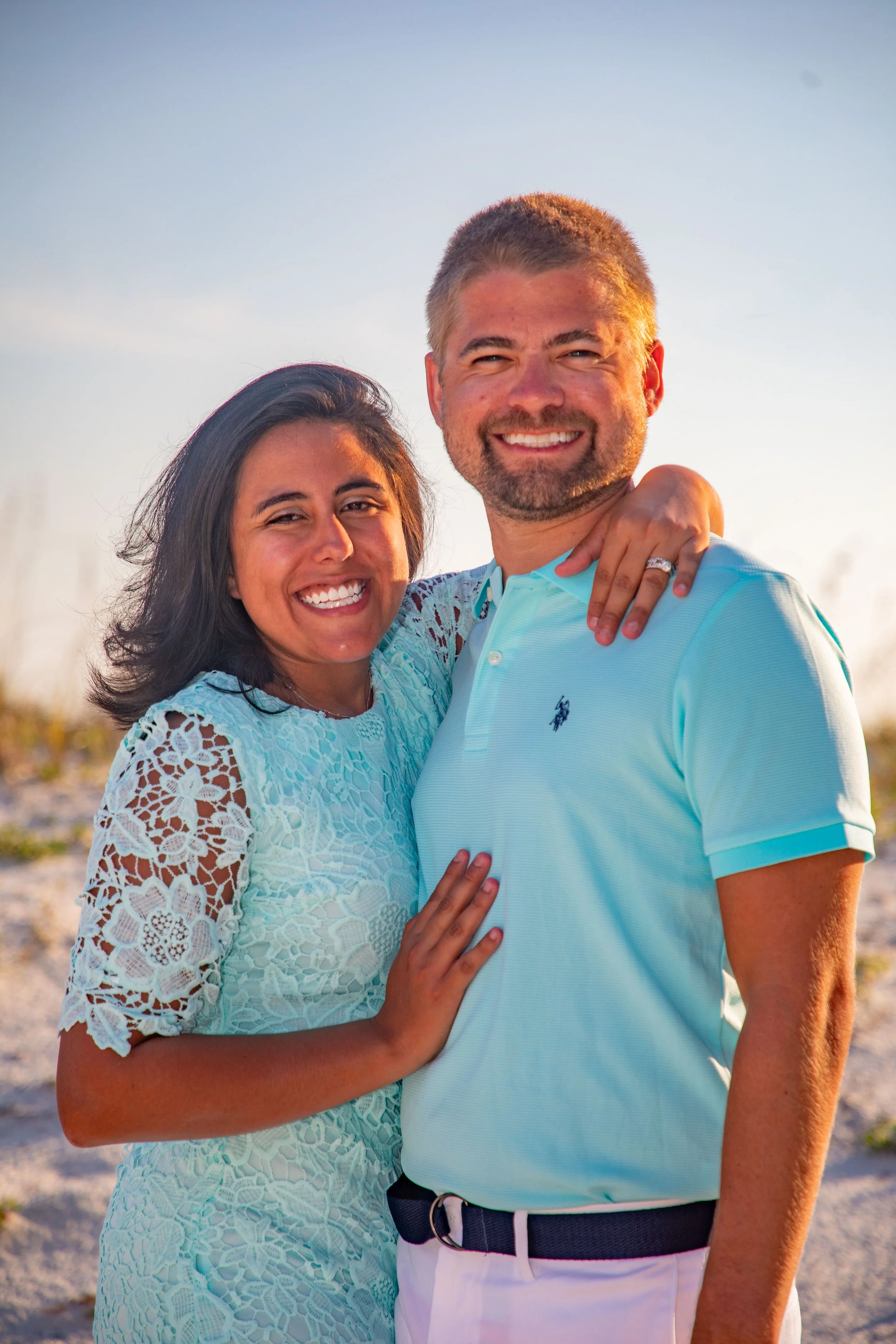 A smiling couple posing outdoors, with the woman wearing a light blue lace dress and the man in a light blue polo shirt. They appear to be on a beach or sandy area.
