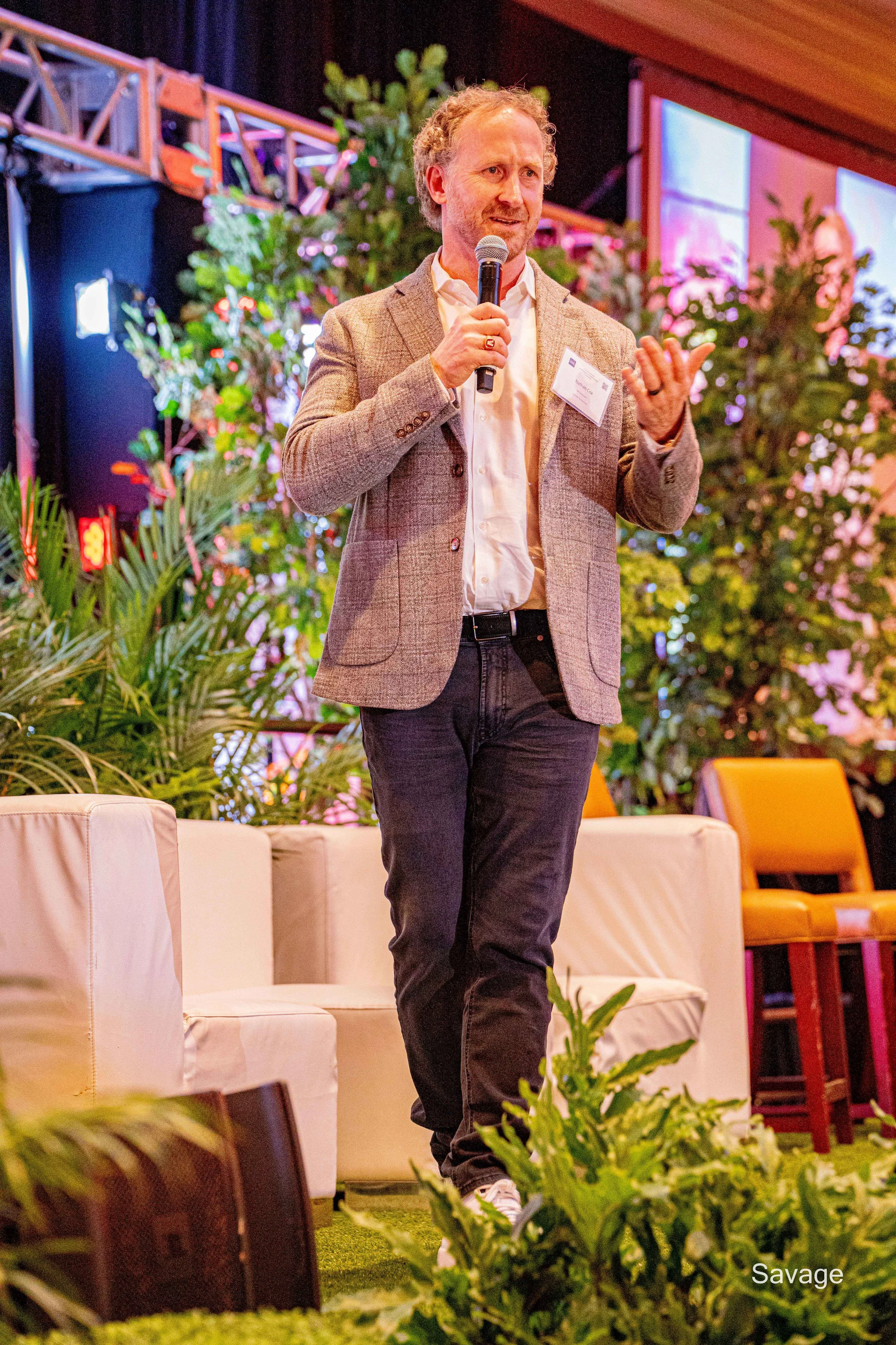 A man speaking into a microphone on stage with plants and a colorful backdrop.