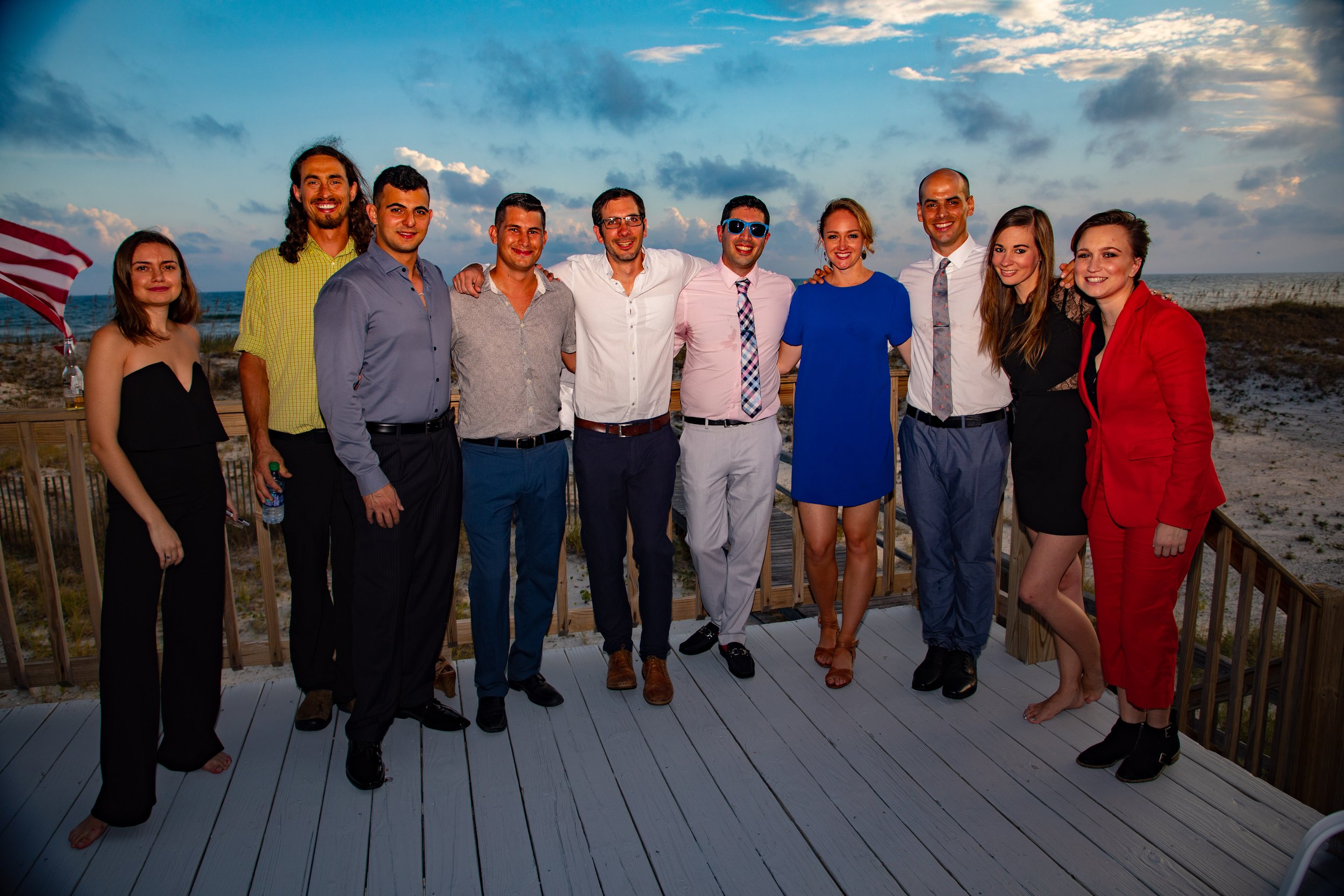 Group of well-dressed people standing on a wooden deck with a beach background.