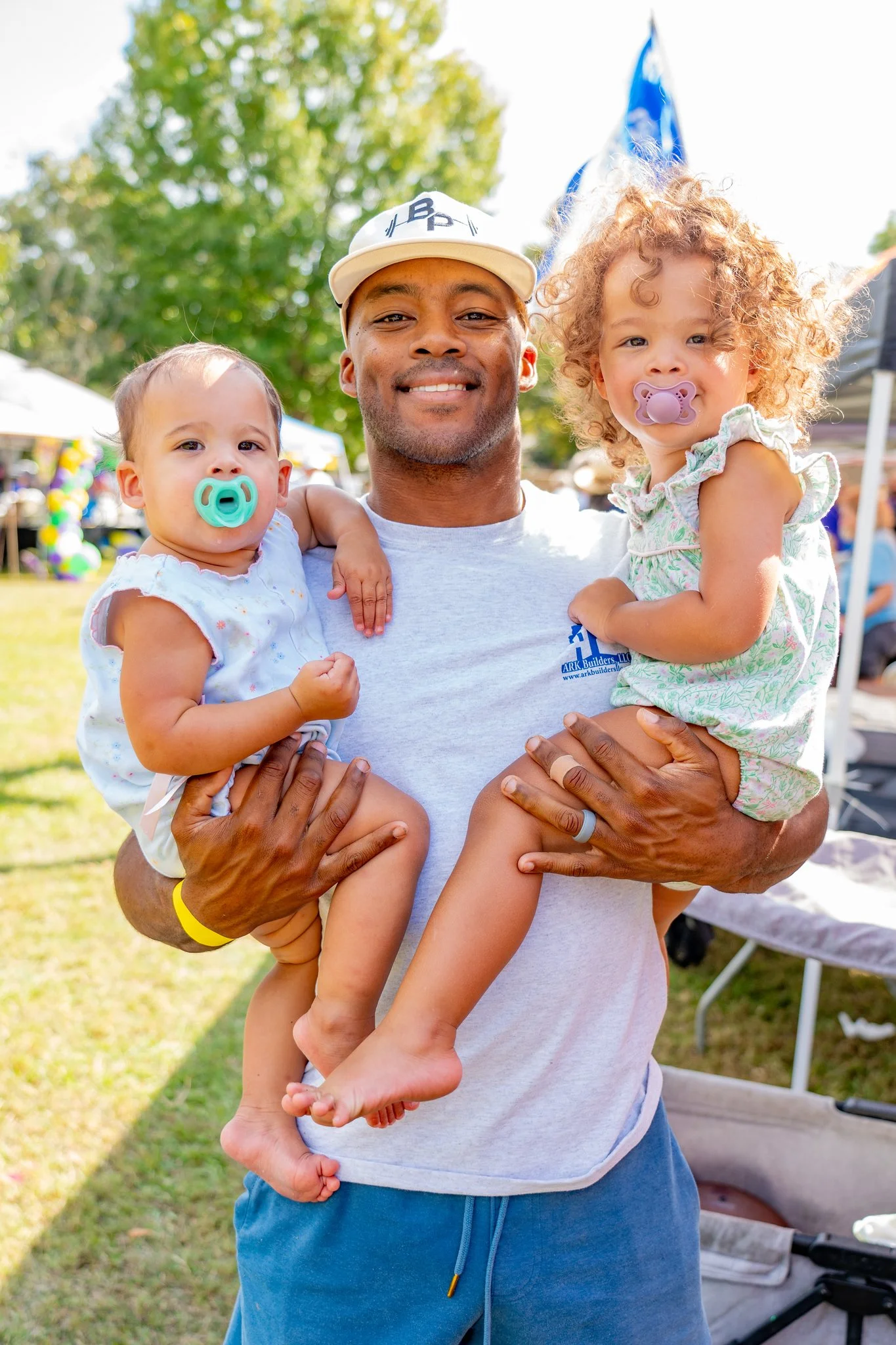 Man smiling holding two young children with pacifiers, outdoor park setting.