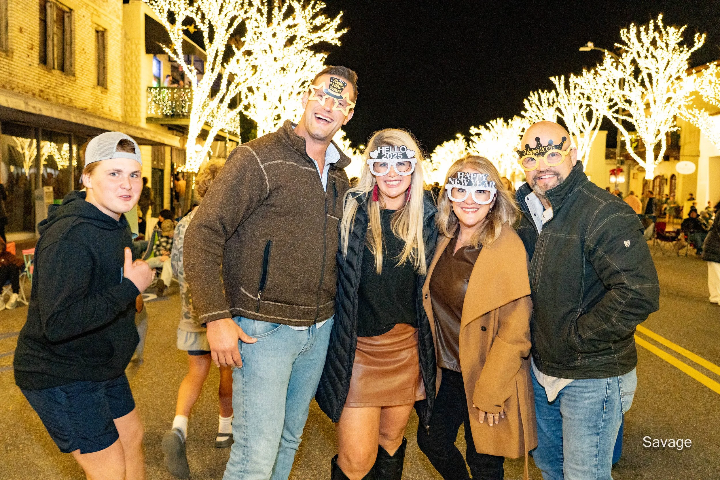 A group of five people posing on a street decorated with bright holiday lights, wearing New Year-themed glasses and smiling.
