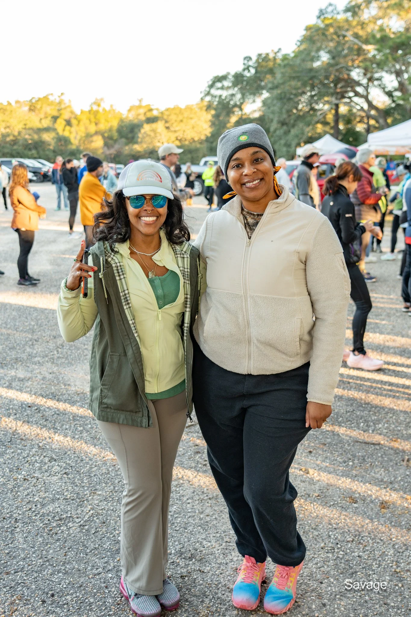 Two women posing outdoors, one wearing a gray beanie and the other wearing a cap and sunglasses, surrounded by people in a park setting with trees and tents in the background.
