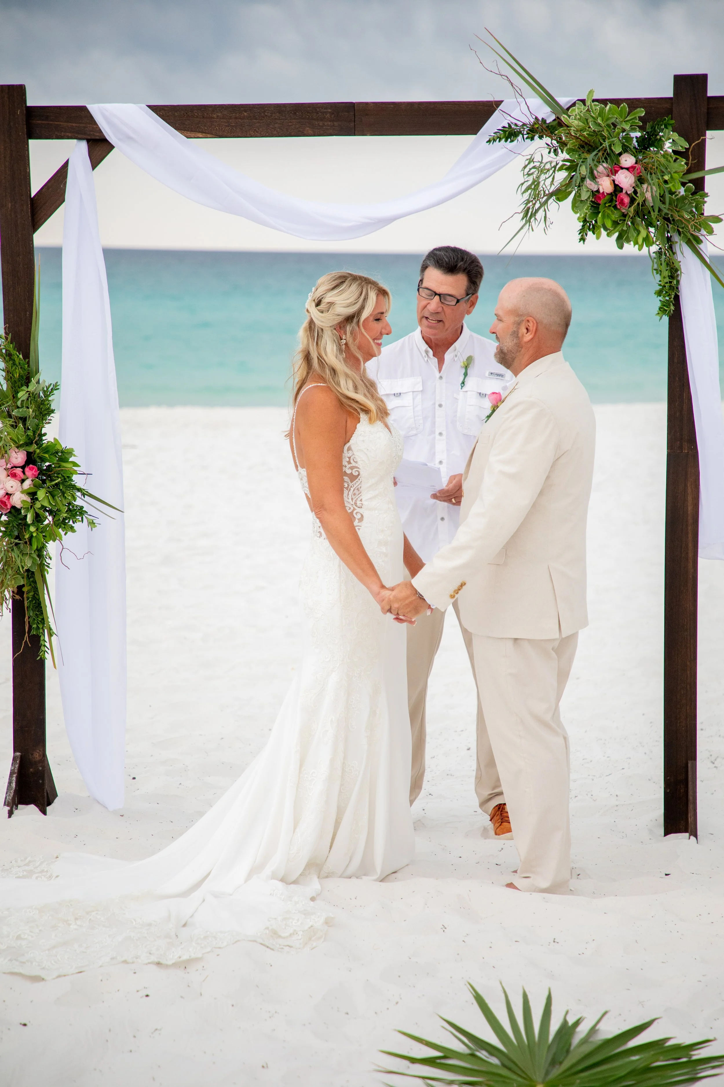 Beach wedding ceremony with a couple holding hands under a decorated arch, officiated by a man in white clothing, with ocean in the background.