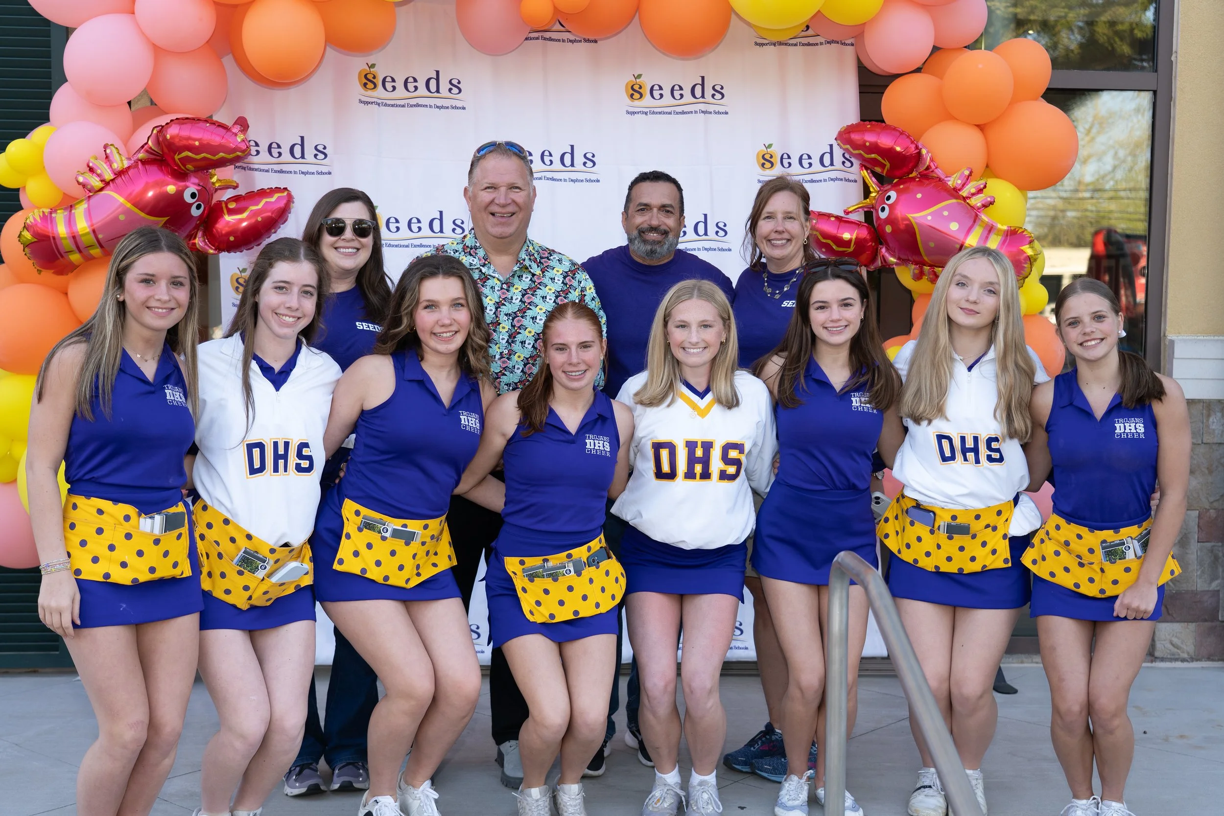 Group of young cheerleaders in blue uniforms with DHS logos, accompanied by three adults, posing in front of a Seeds-branded backdrop and pink and orange balloon arch.