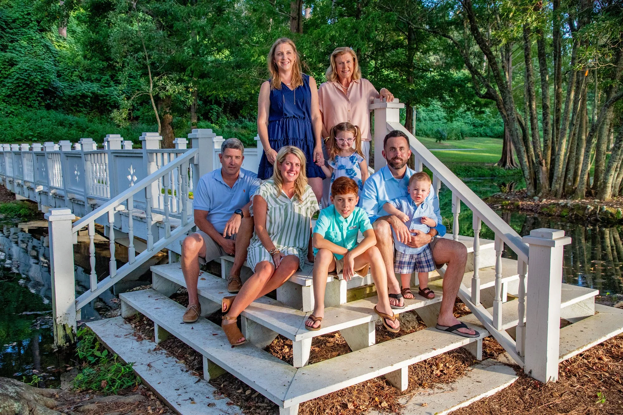 Family posing on steps near a white bridge in a park, surrounded by greenery.