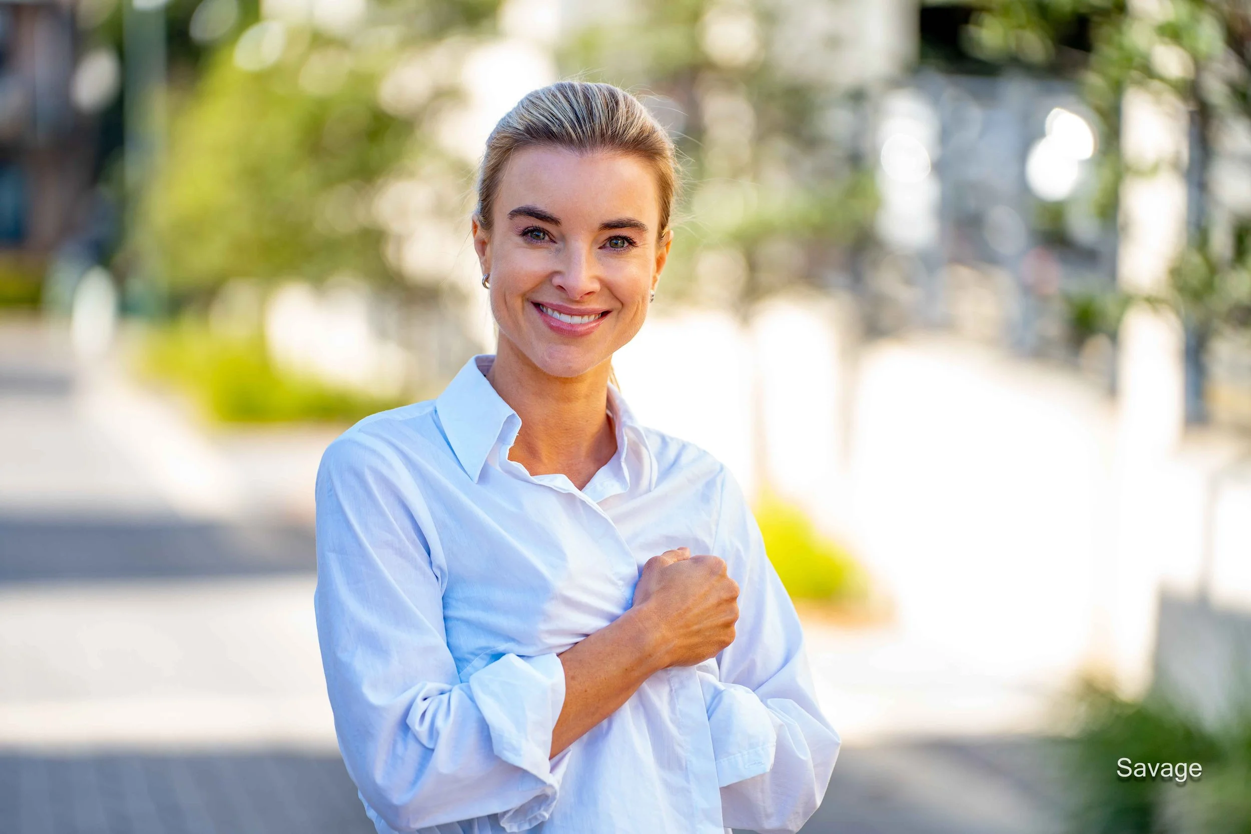 A smiling person in a white shirt stands outdoors on a sunny street, surrounded by blurred greenery.