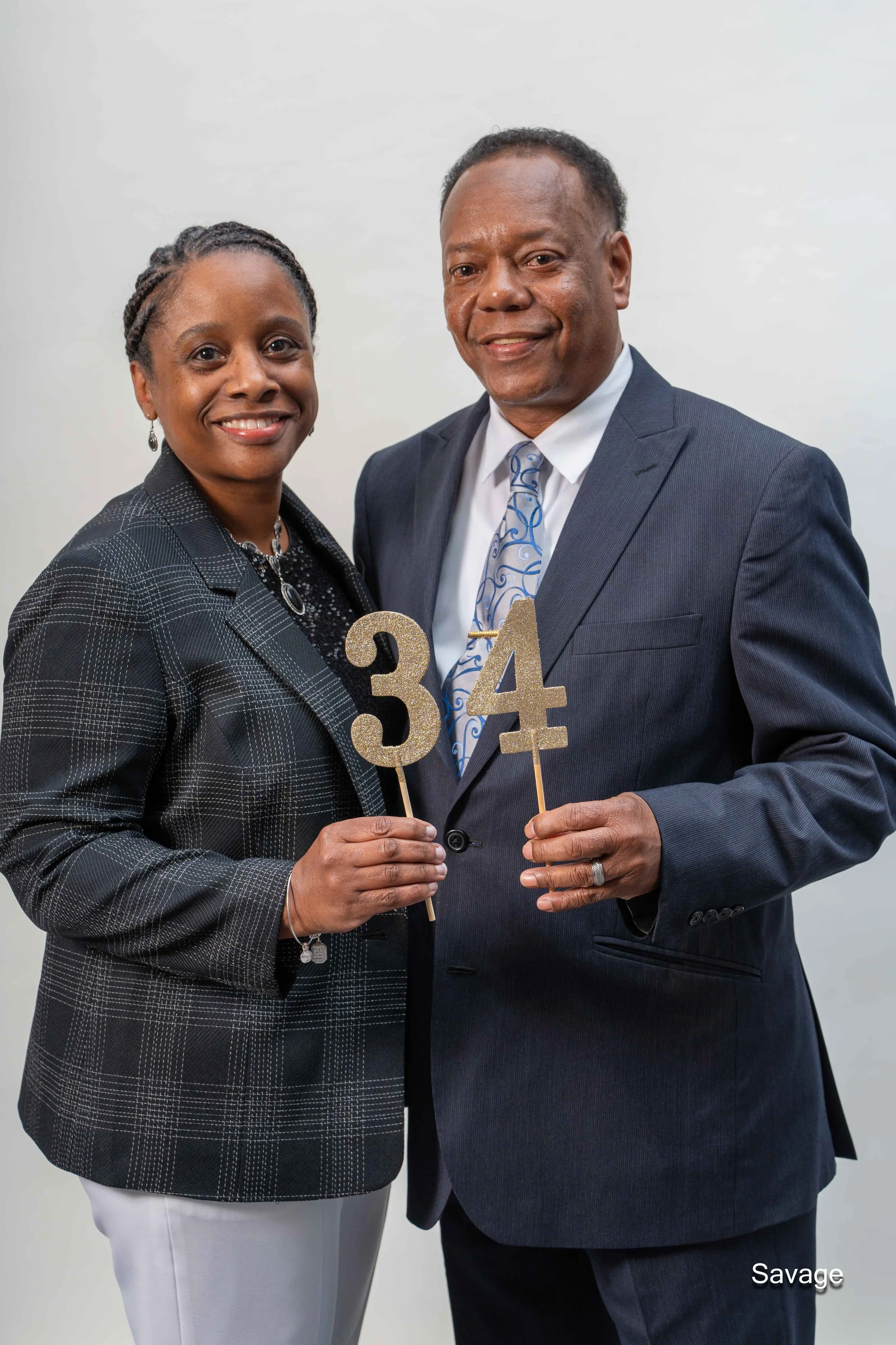 A smiling couple in formal attire holding gold number cutouts "34" against a plain background. The woman is wearing a plaid jacket and the man is in a suit and tie.