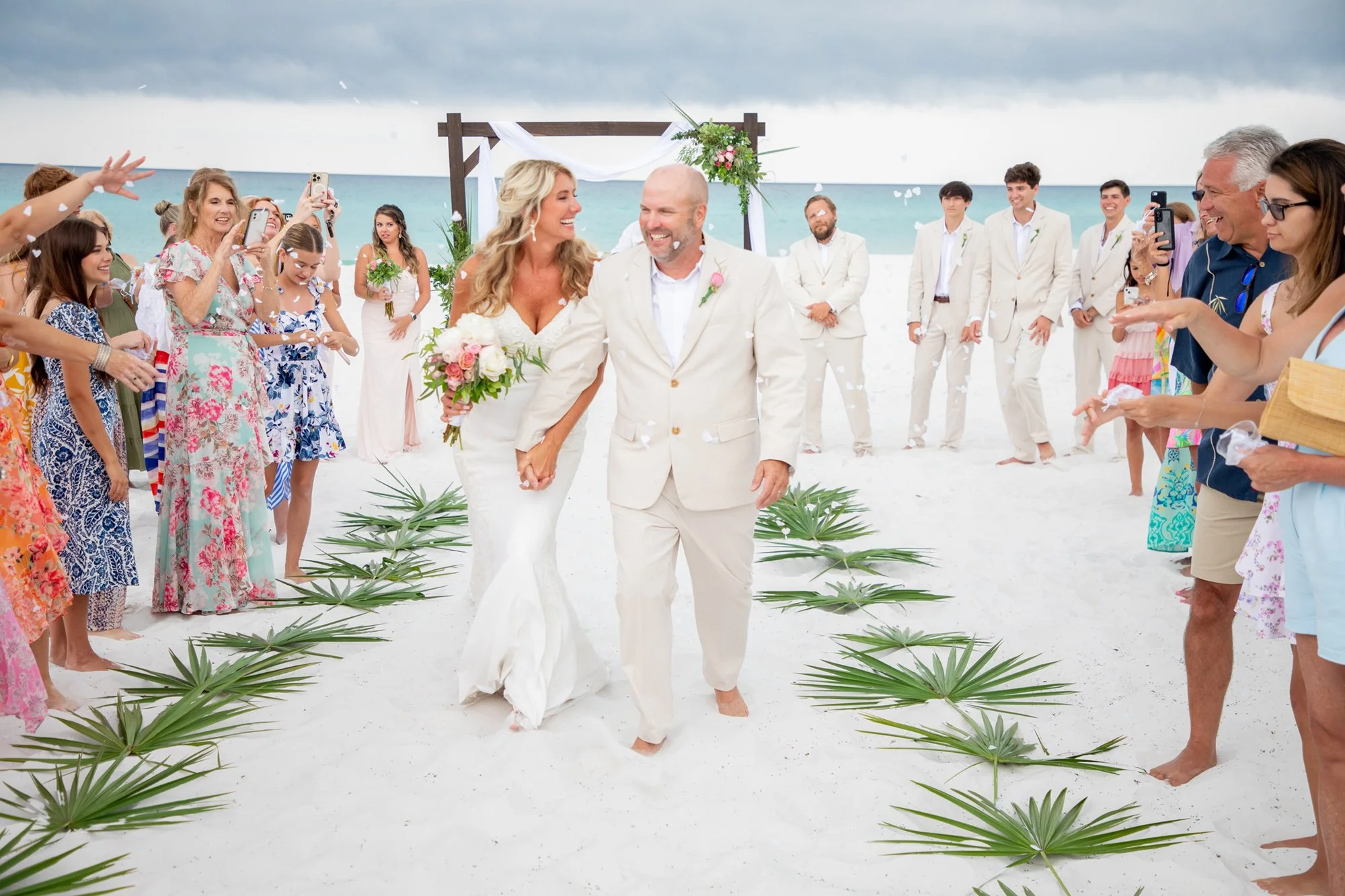 A bride and groom in white attire walk down a sandy aisle on a beach, surrounded by smiling guests and greenery. The ocean is visible in the background.