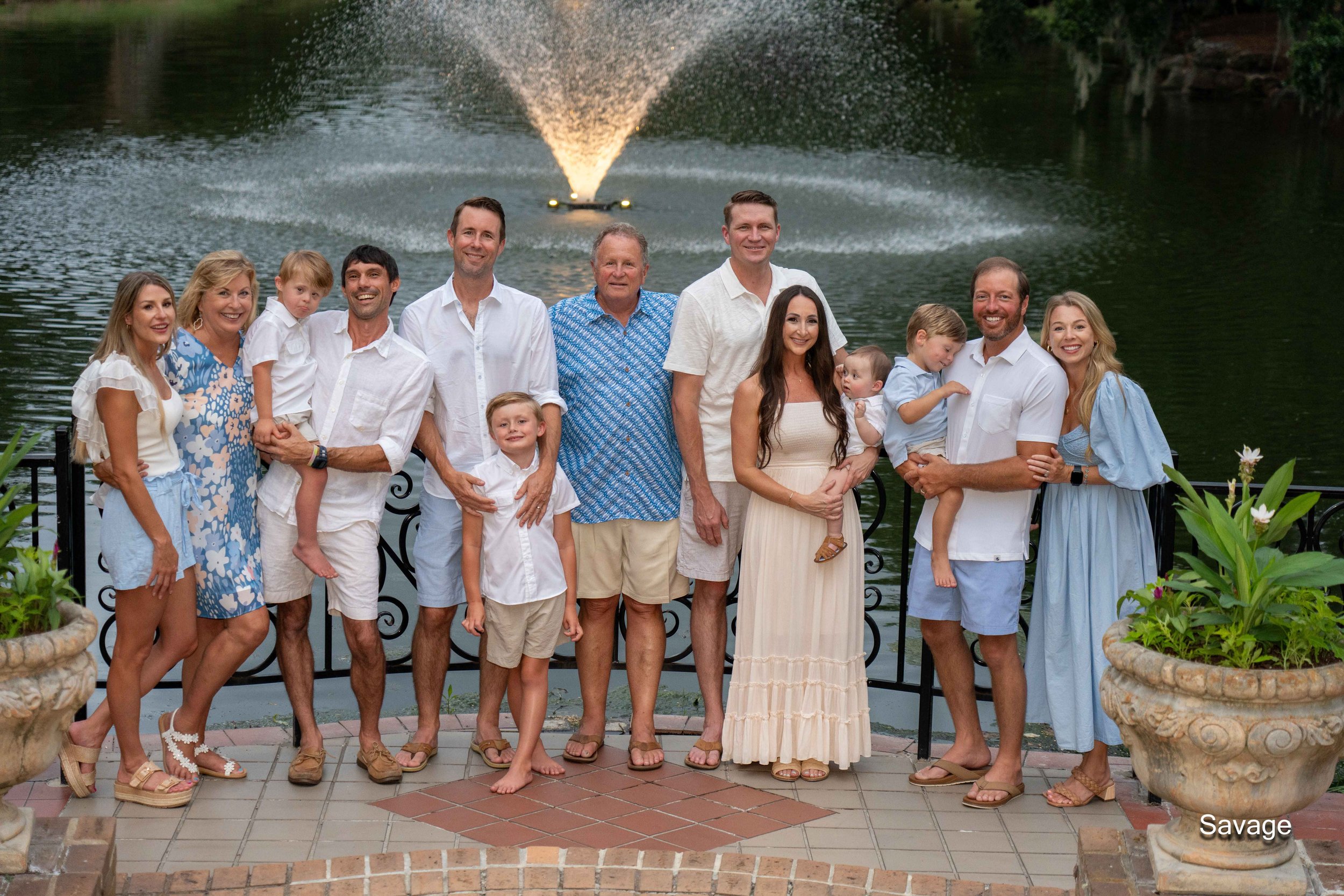 Group of people posing by a lake with a fountain, wearing summer attire. Includes adults and children, with greenery and potted plants in the foreground.