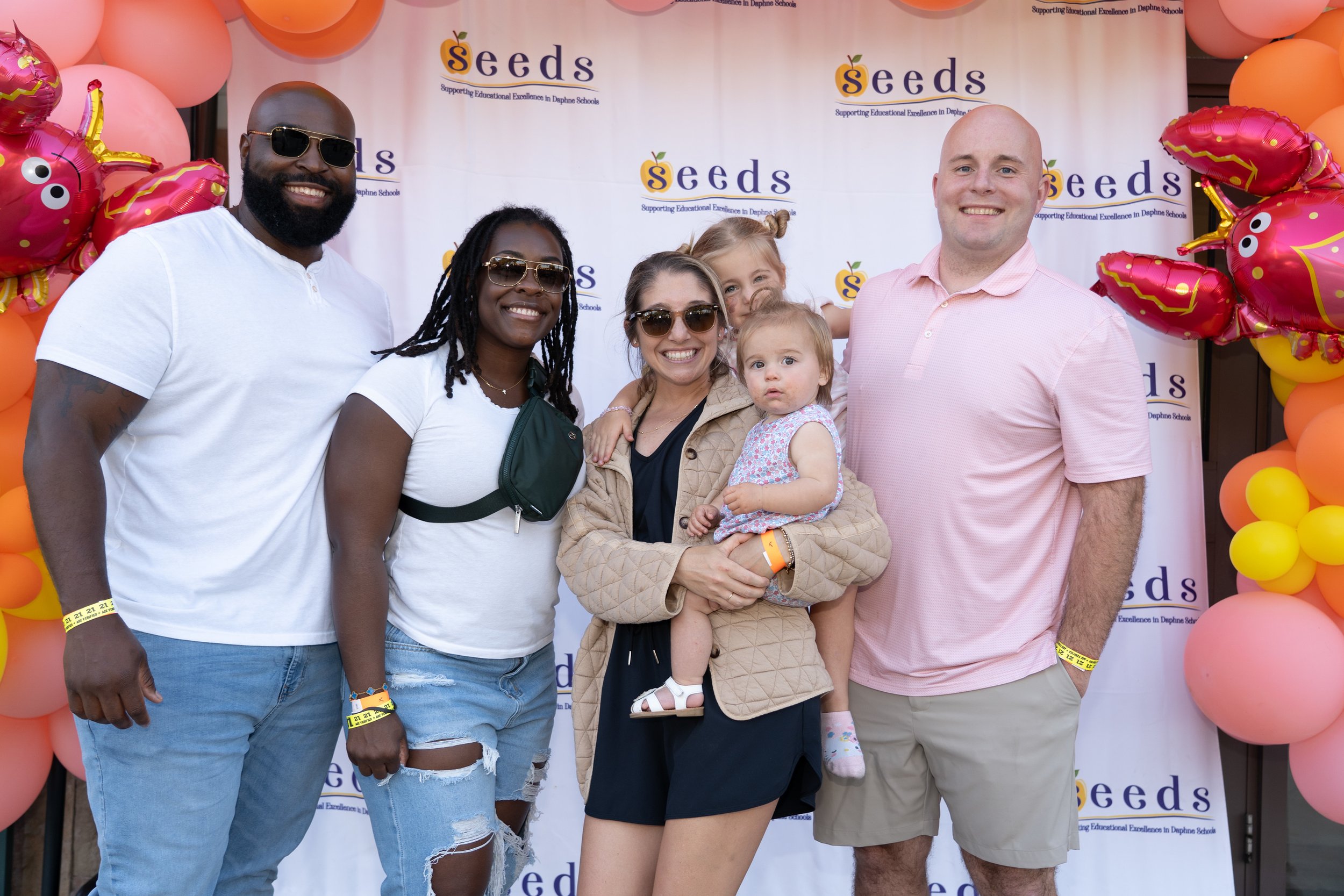Group of adults and children smiling at an event with colorful balloon decorations and a banner that says "Seeds."