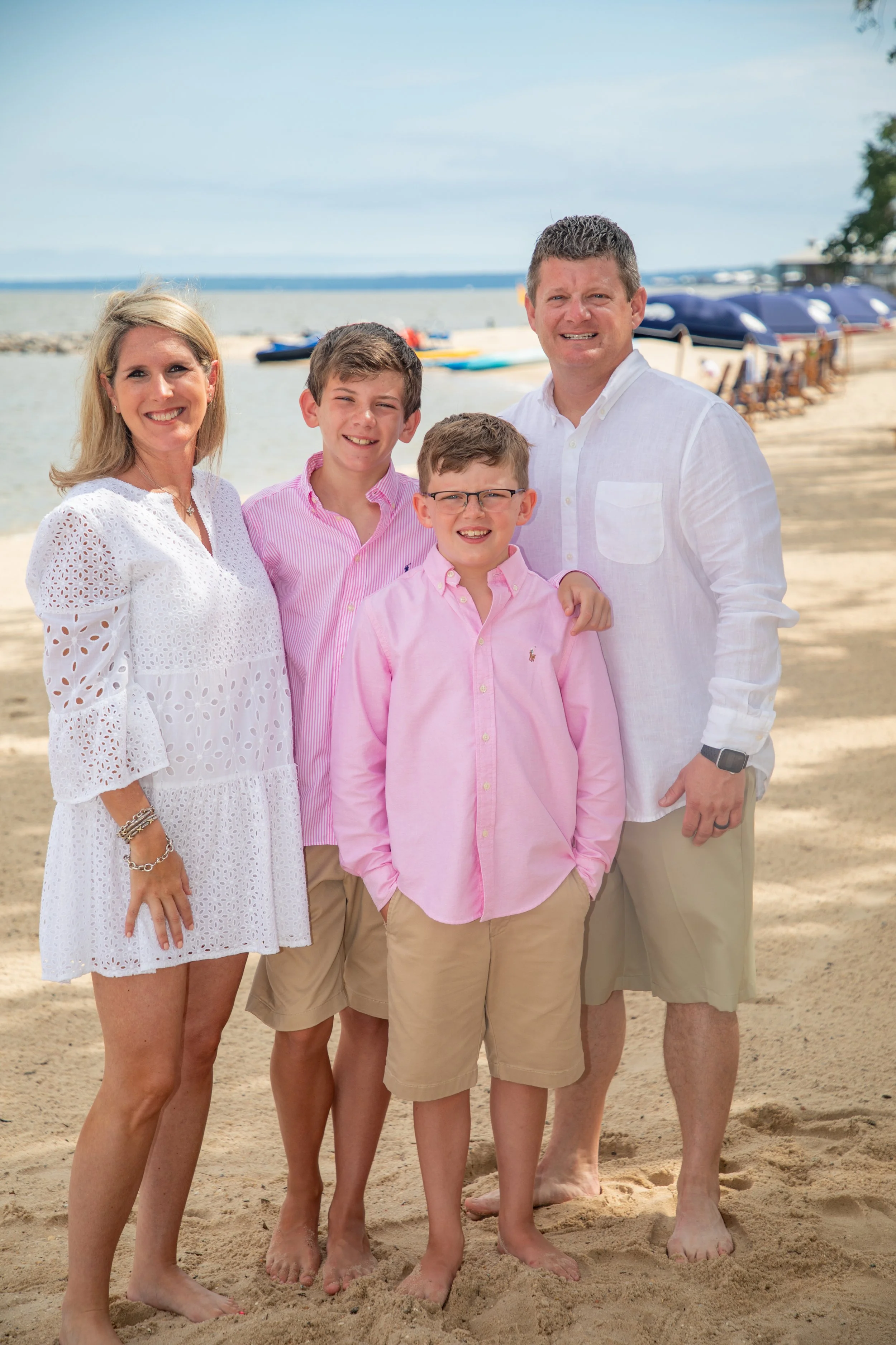 A family of four posing together on a sandy beach, with the ocean and beach chairs in the background. The two boys are wearing pink shirts and khaki shorts, while the woman is in a white dress and the man is in a white shirt and khaki shorts.
