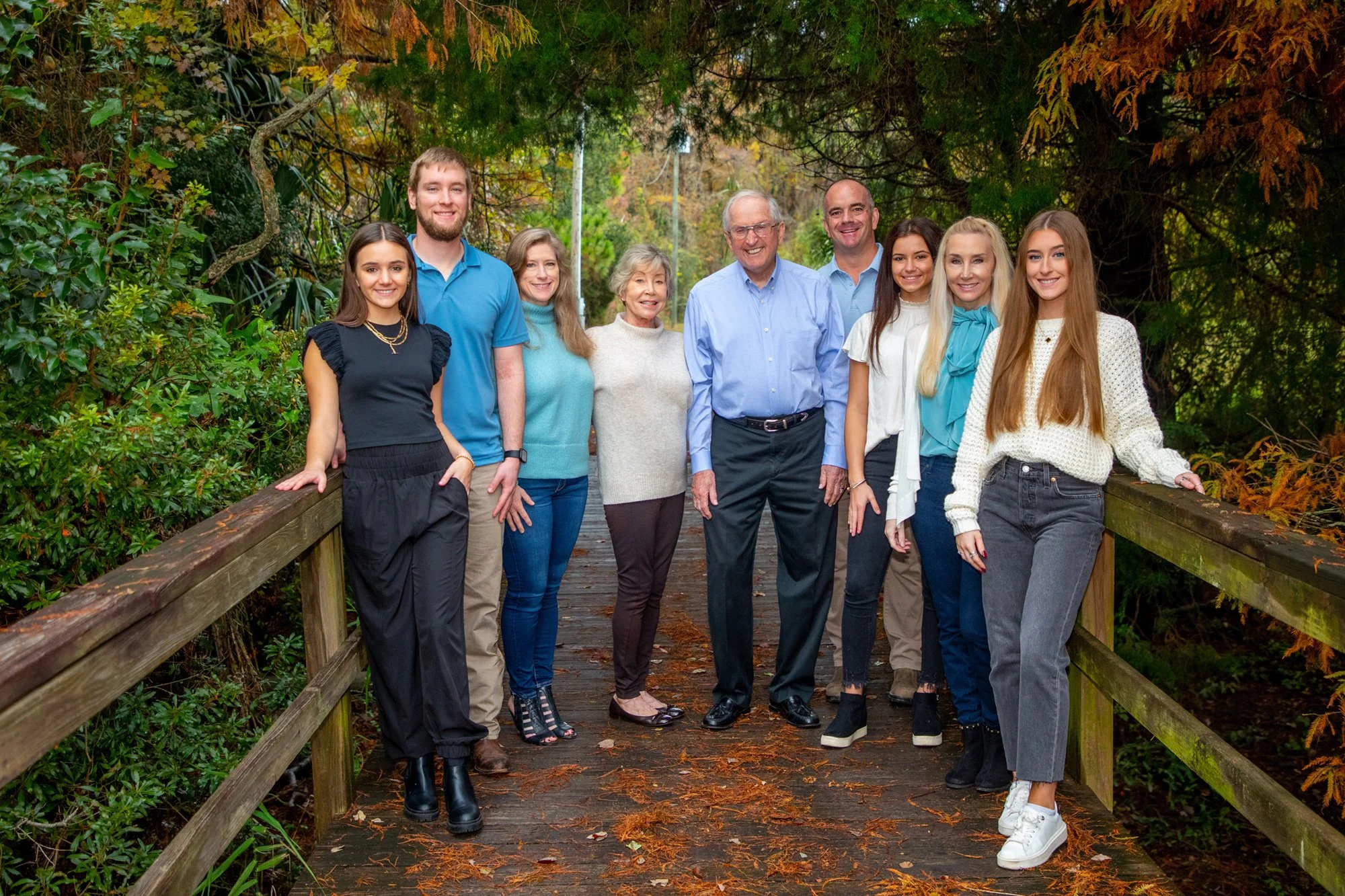 Group of eight people standing on a wooden bridge surrounded by greenery and autumn foliage, smiling at the camera.